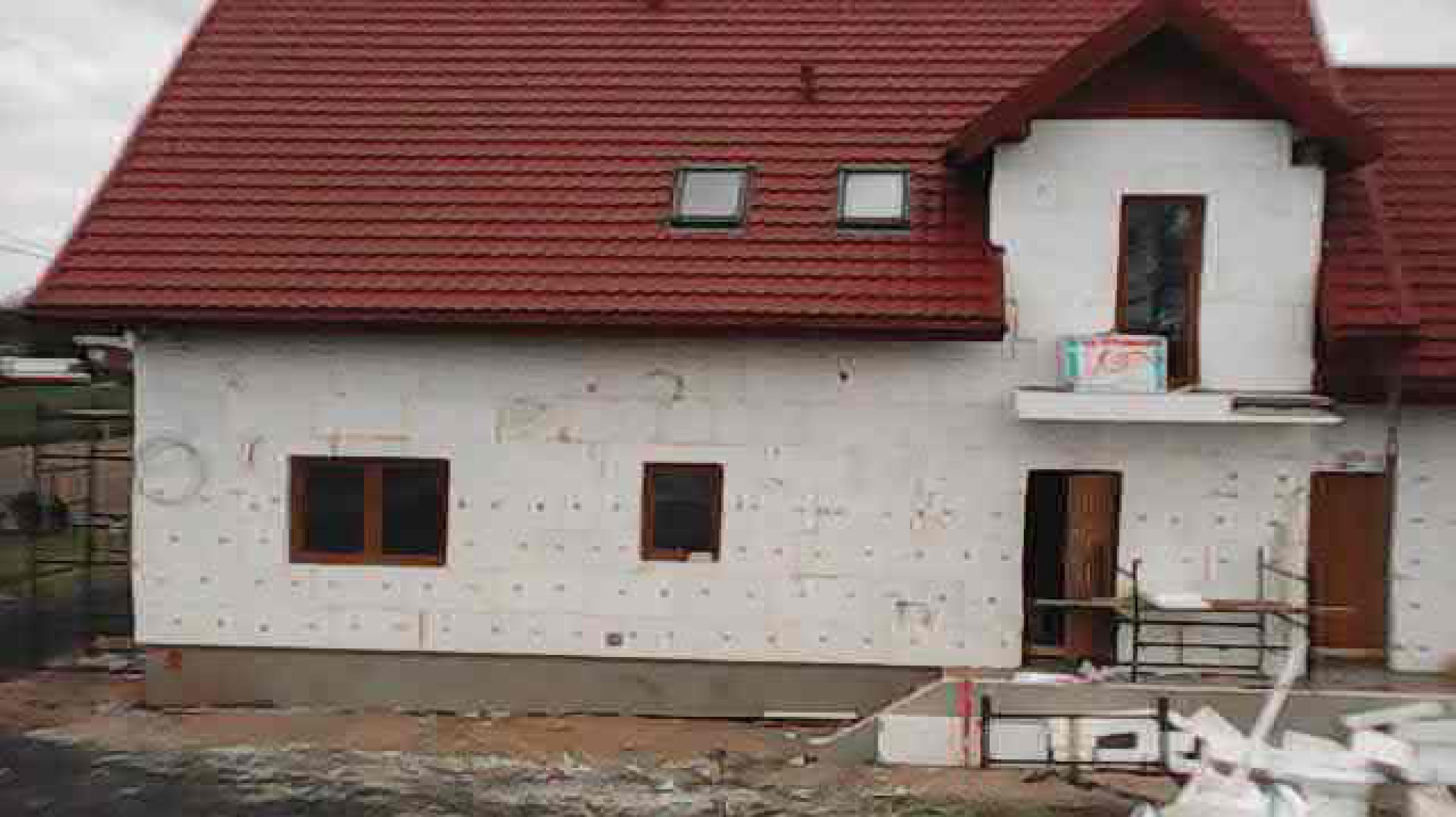 Dom w trakcie ocieplania: czerwony dach, biała elewacja z widoczną izolacją, okna i drzwi. Na dole widoczne materiały budowlane i rusztowanie.