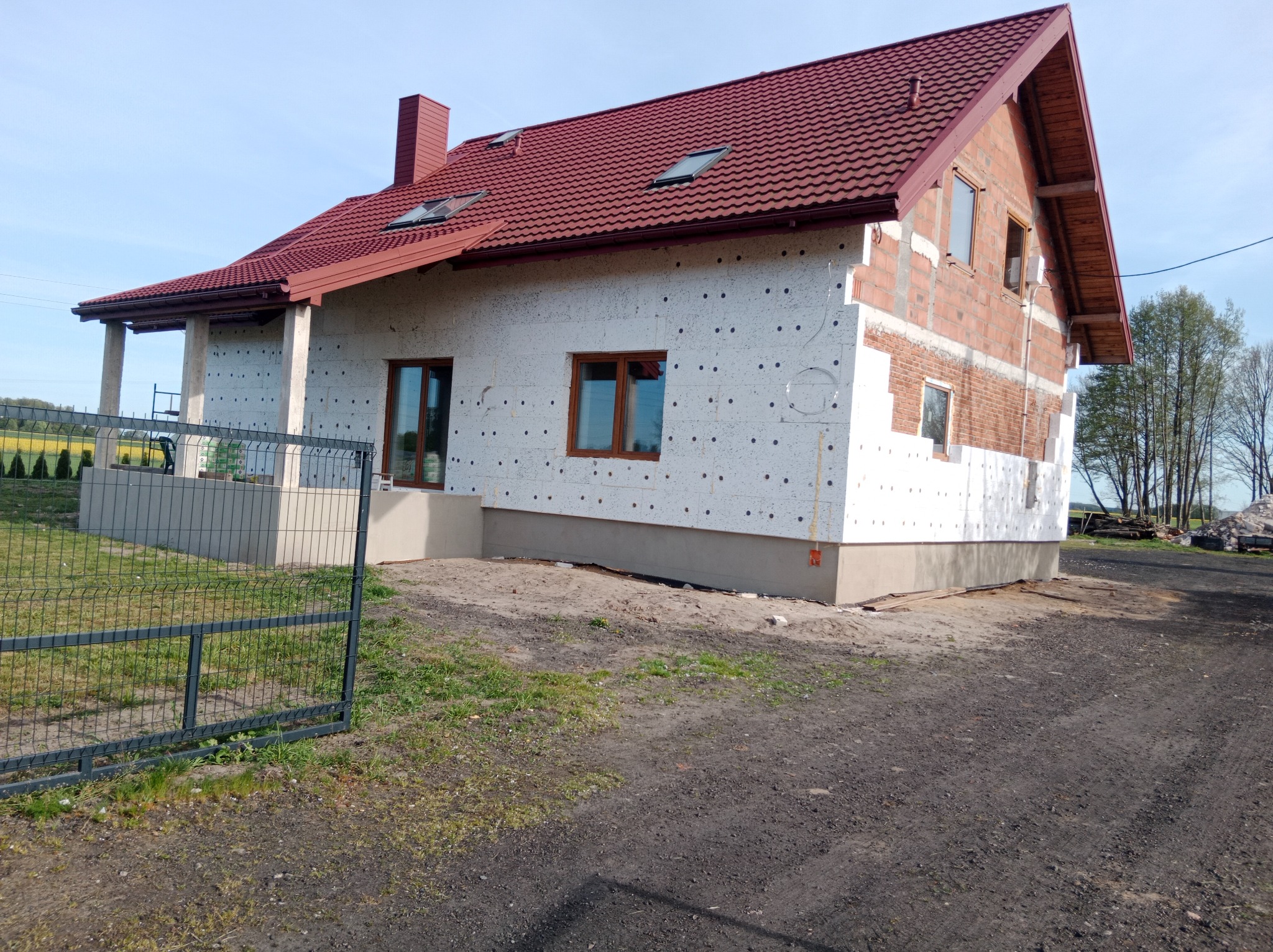 Dom jednorodzinny w trakcie ocieplania elewacji styropianem, widok z zewnątrz, częściowo otynkowany, z czerwoną dachówką i oknami z brązowymi ramami.