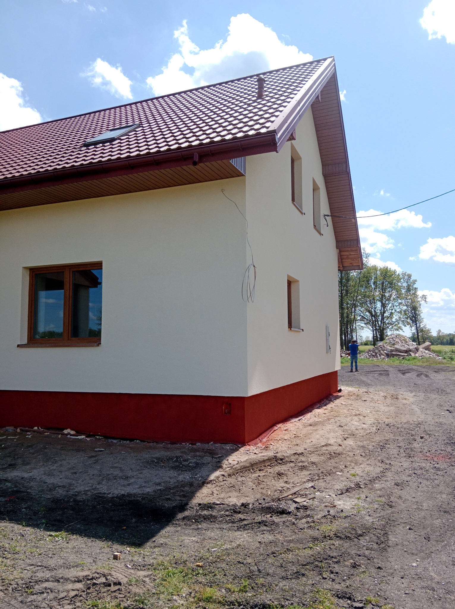 Świeżo otynkowany, dwukondygnacyjny dom z brązowym dachem i oknami, z czerwonym cokołem, na tle błękitnego nieba i zieleni. Widoczne niedociągnięcia.