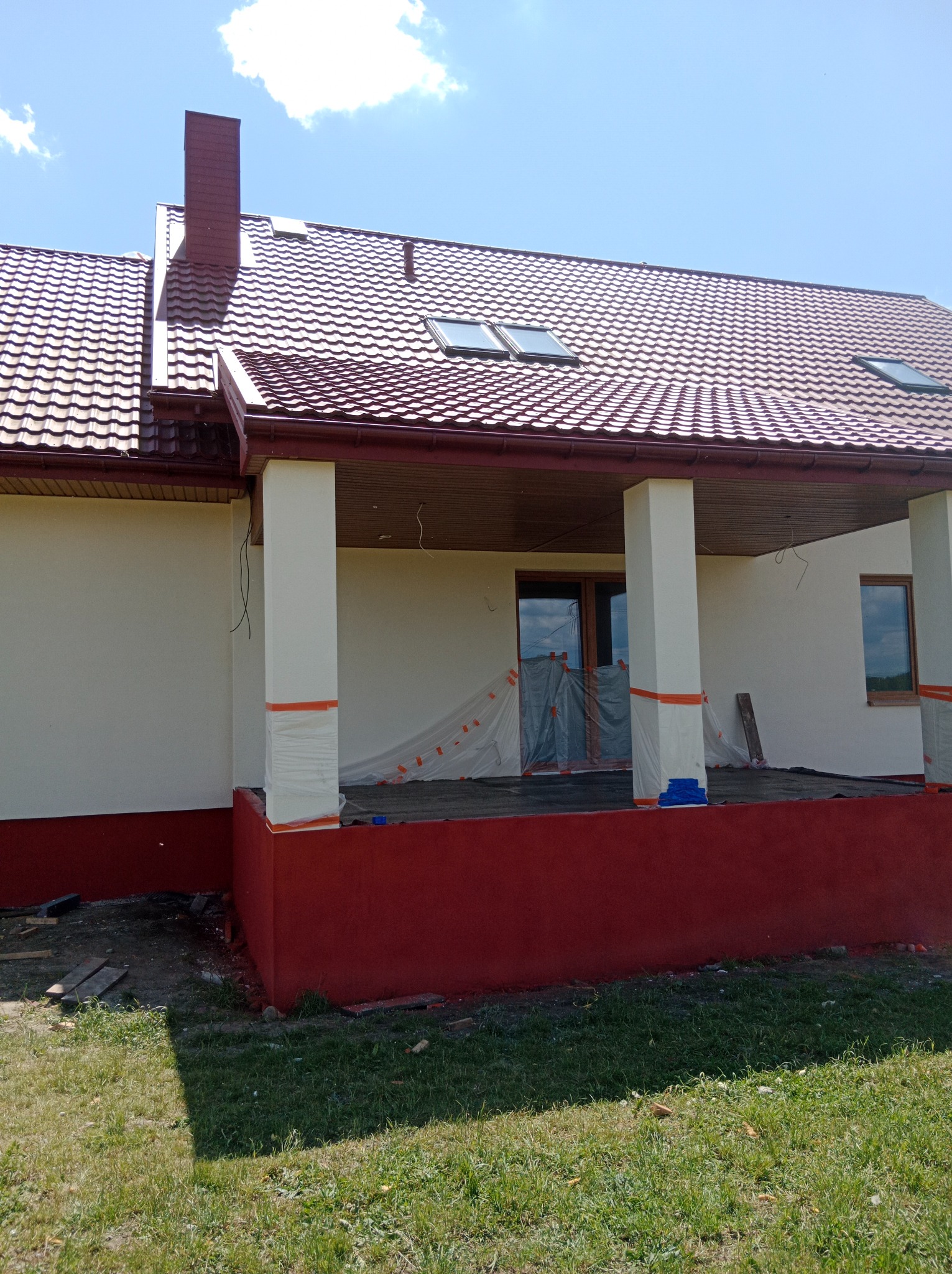 Dom jednorodzinny w trakcie prac wykończeniowych elewacji: malowanie ścian na kremowo i czerwono, zabezpieczone okna, widoczny dach z brązowej blachy.