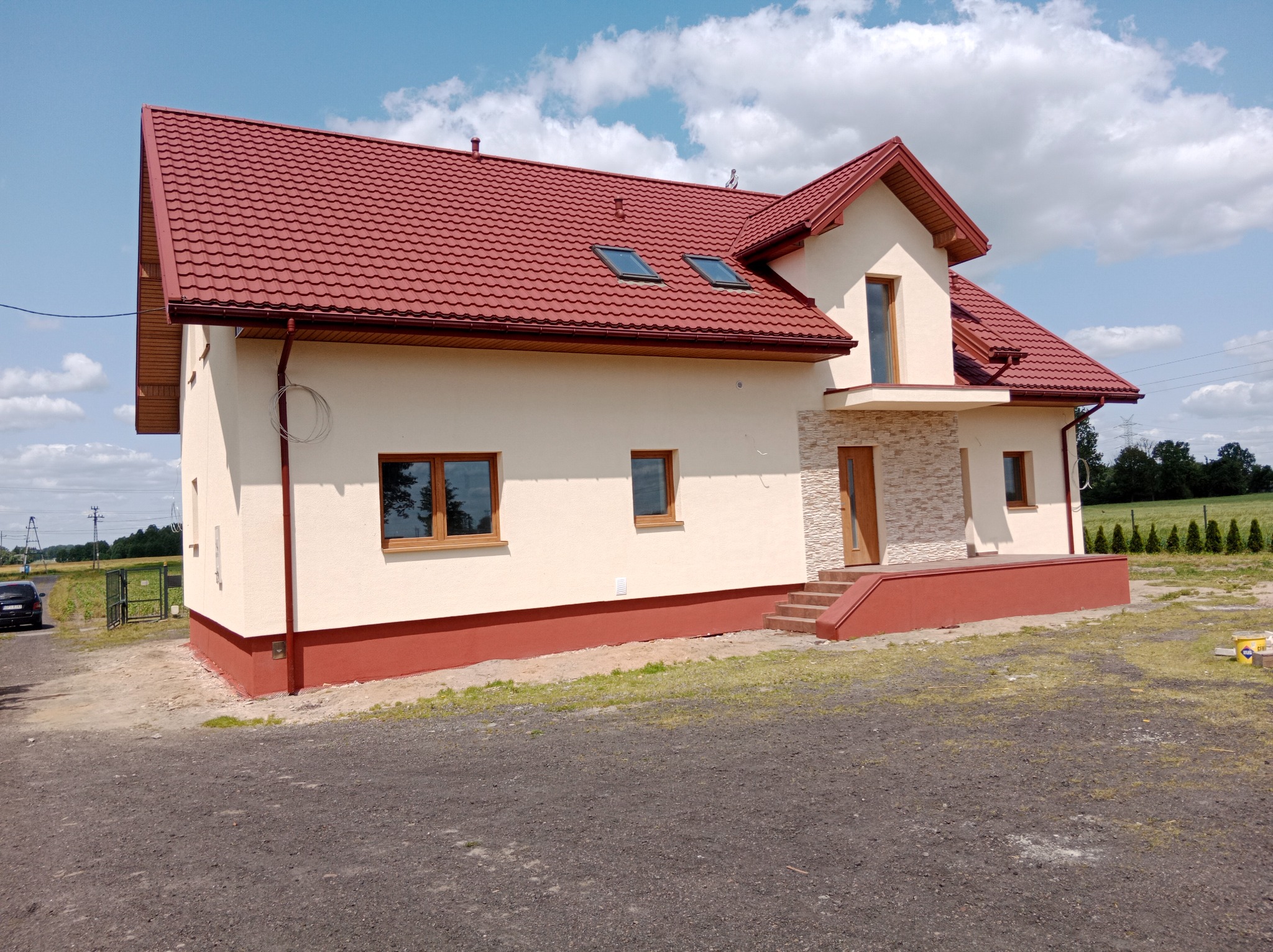 Elewacja domu jednorodzinnego z czerwonym dachem, kremową fasadą i kamiennym wykończeniem wejścia. Widoczne okna z drewnianą ramą i fragment otoczenia.