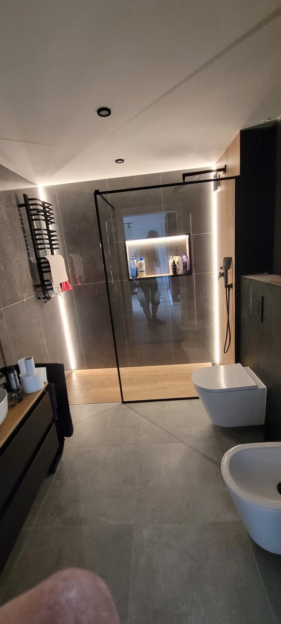 Nowoczesna łazienka z kabiną typu walk-in, szarymi płytkami i podświetleniem LED. Minimalistyczny design z wiszącą toaletą i bidetem. Widoczny grzejnik drabinkowy.