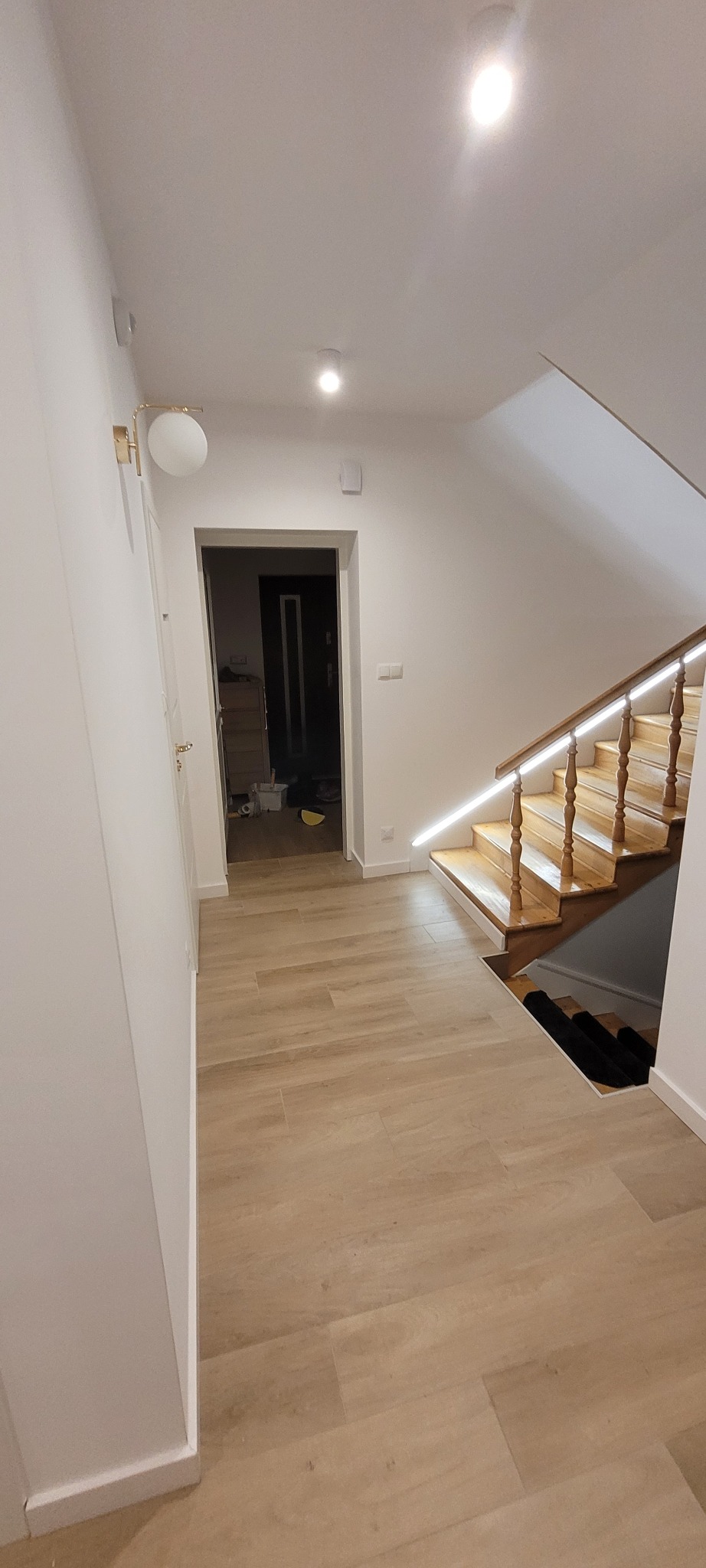 Jasne płytki podłogowe imitujące drewno w korytarzu z drewnianymi schodami z podświetleniem LED. Widoczne drzwi i lampa ścienna.