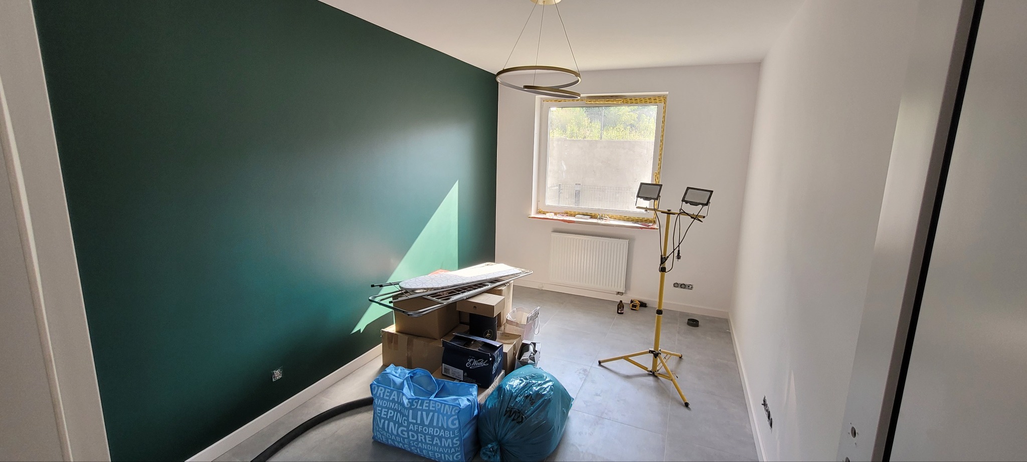 Pokój po malowaniu z jedną ścianą w kolorze butelkowej zieleni. Widoczne kartony, worki i lampa budowlana. Okno z grzejnikiem. Nowoczesna lampa sufitowa.