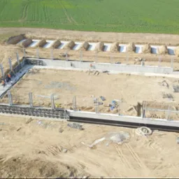 Widok z drona na plac budowy z wykopanymi fundamentami, częściowo wylane ściany fundamentowe oraz widoczne szalunki i zbrojenia. W tle zielone pole.