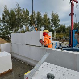 Wznoszenie ściany z betonu komórkowego Ytong. Robotnik w kasku i odzieży roboczej układa bloczki za pomocą dźwigu. Widoczne narzędzia i elementy konstrukcyjne.