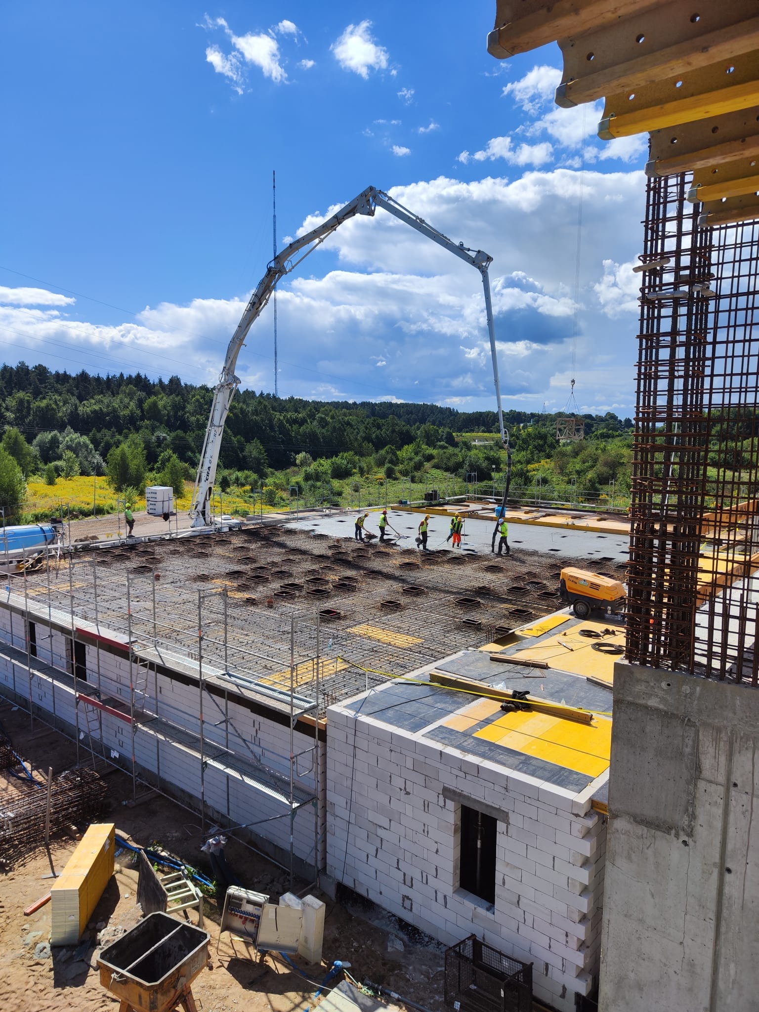 Betonowanie stropu budynku w trakcie budowy, widoczna pompa do betonu, pracownicy w kaskach i kamizelkach odblaskowych, zbrojenie i szalunki, w tle las i błękitne niebo z chmurami.