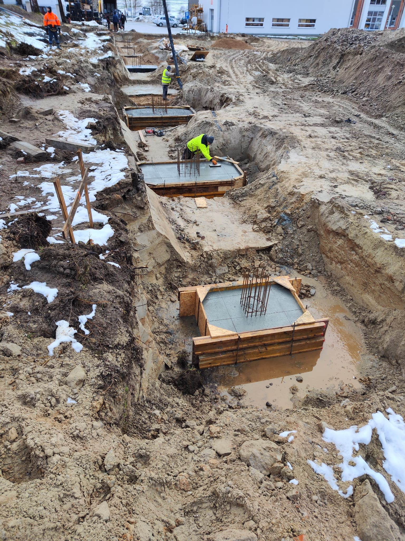 Wykop pod fundamenty z drewnianym szalunkiem i zbrojeniem, częściowo zalany wodą, w tle pracownicy przy pompie do betonu.