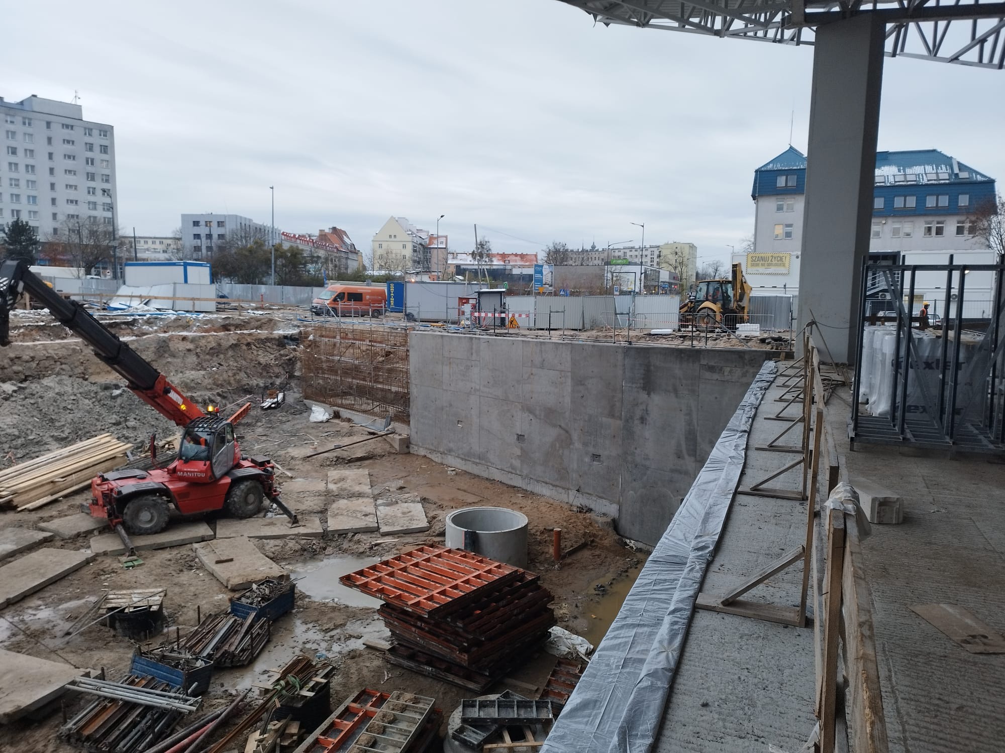 Widok z góry na plac budowy z betonową ścianą fundamentową, stosem pomarańczowych szalunków, czerwonym wózkiem widłowym Manitou i koparką w tle, w pochmurny dzień.