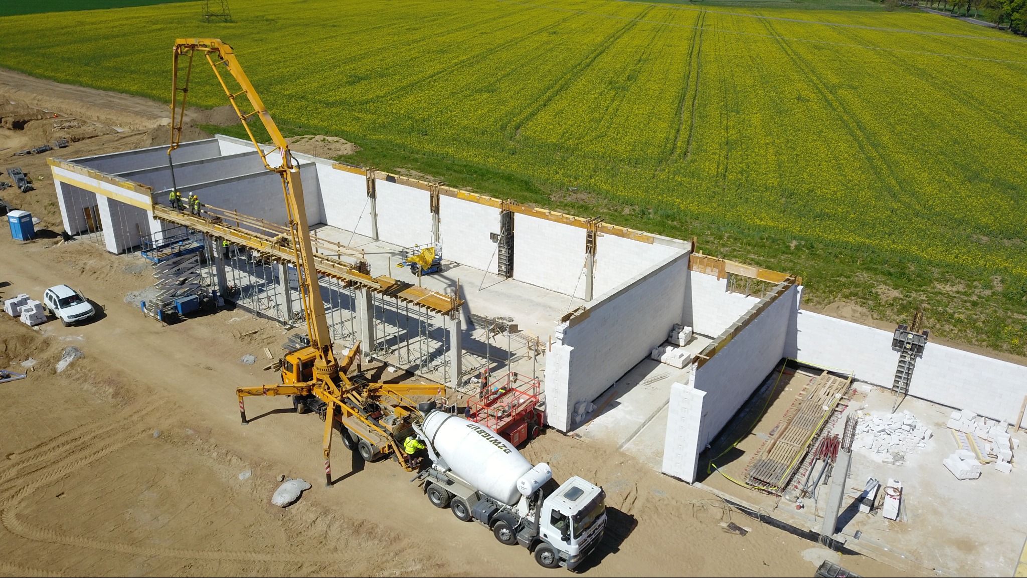 Wysoka perspektywa: budowa budynku z betonu komórkowego w trakcie wylewania, z użyciem pompy do betonu na tle pola rzepaku w słoneczny dzień.