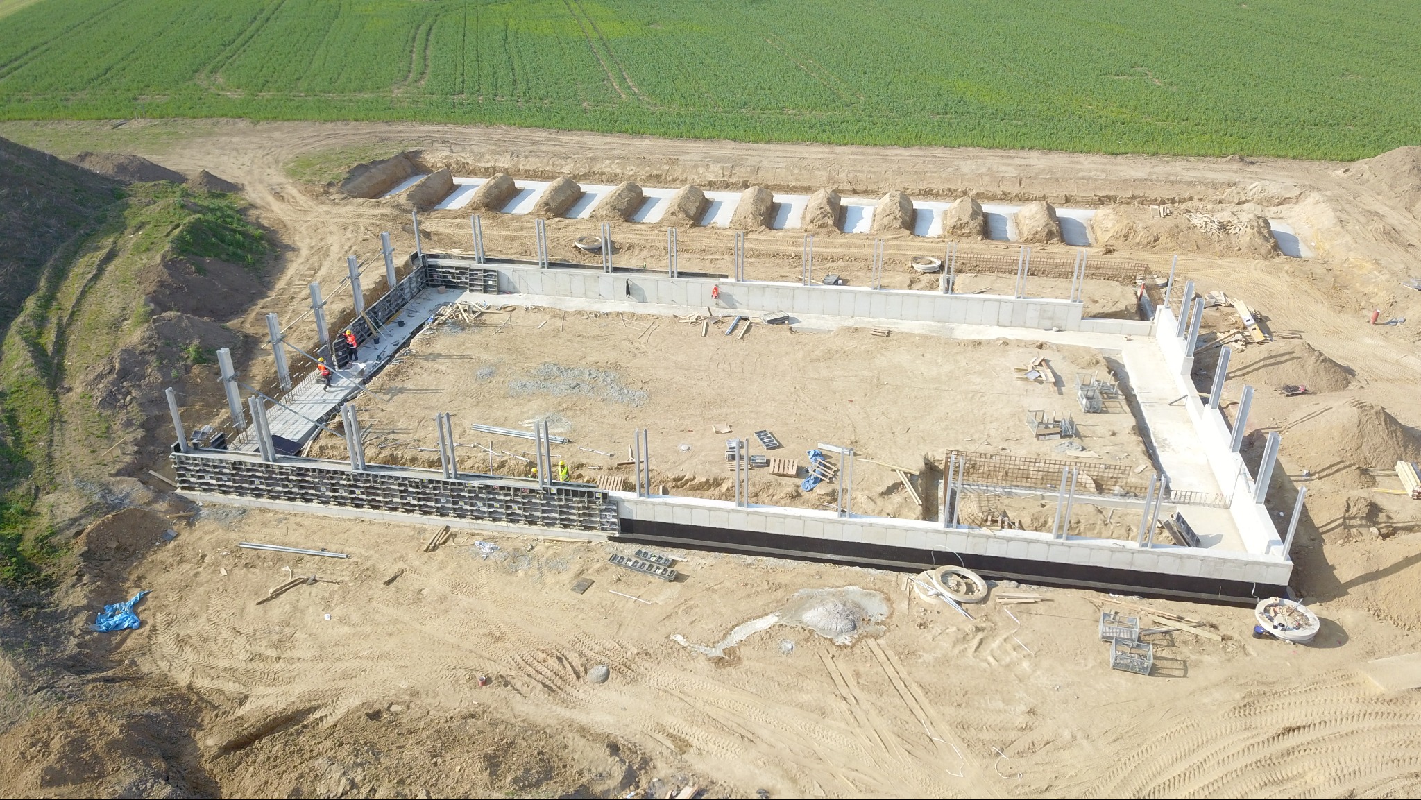 Widok z drona na plac budowy z wykopanymi fundamentami, częściowo wylane ściany fundamentowe oraz widoczne szalunki i zbrojenia. W tle zielone pole.