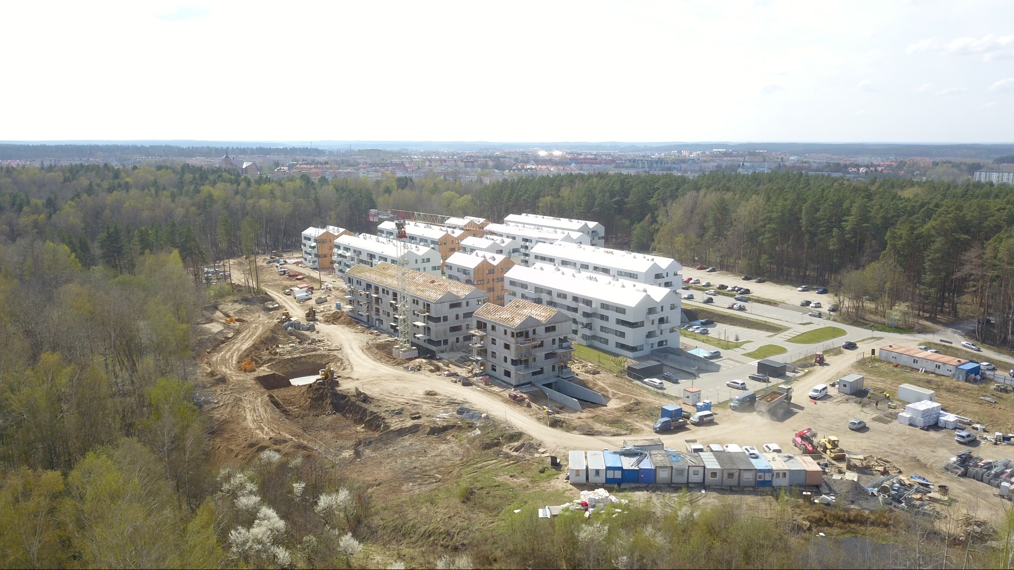 Widok z drona na plac budowy osiedla z budynkami w różnym stopniu ukończenia, otoczonego lasem i miastem w tle. Na pierwszym planie wykop i ciężki sprzęt.