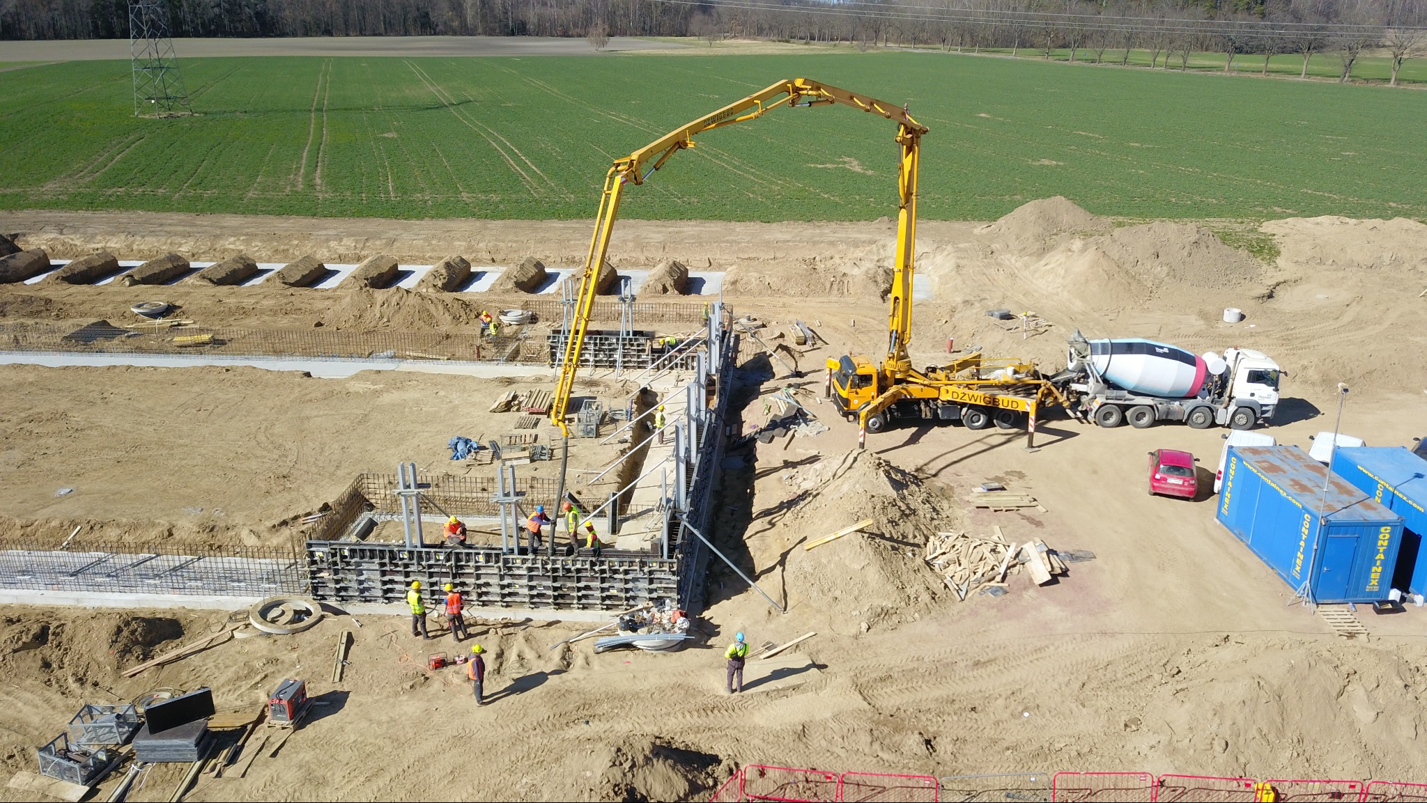 Betonowanie fundamentów szalunkowych z użyciem pompy do betonu. Widoczni pracownicy w kaskach i kamizelkach odblaskowych. W tle pole uprawne i las.
