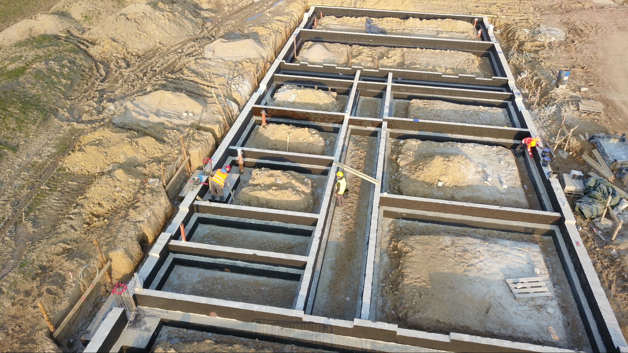 Z góry widok na fundamenty domu z cegły, zabezpieczone czarną izolacją. Dwóch pracowników w kamizelkach wykonuje prace. Wokół ziemia i ślady opon.