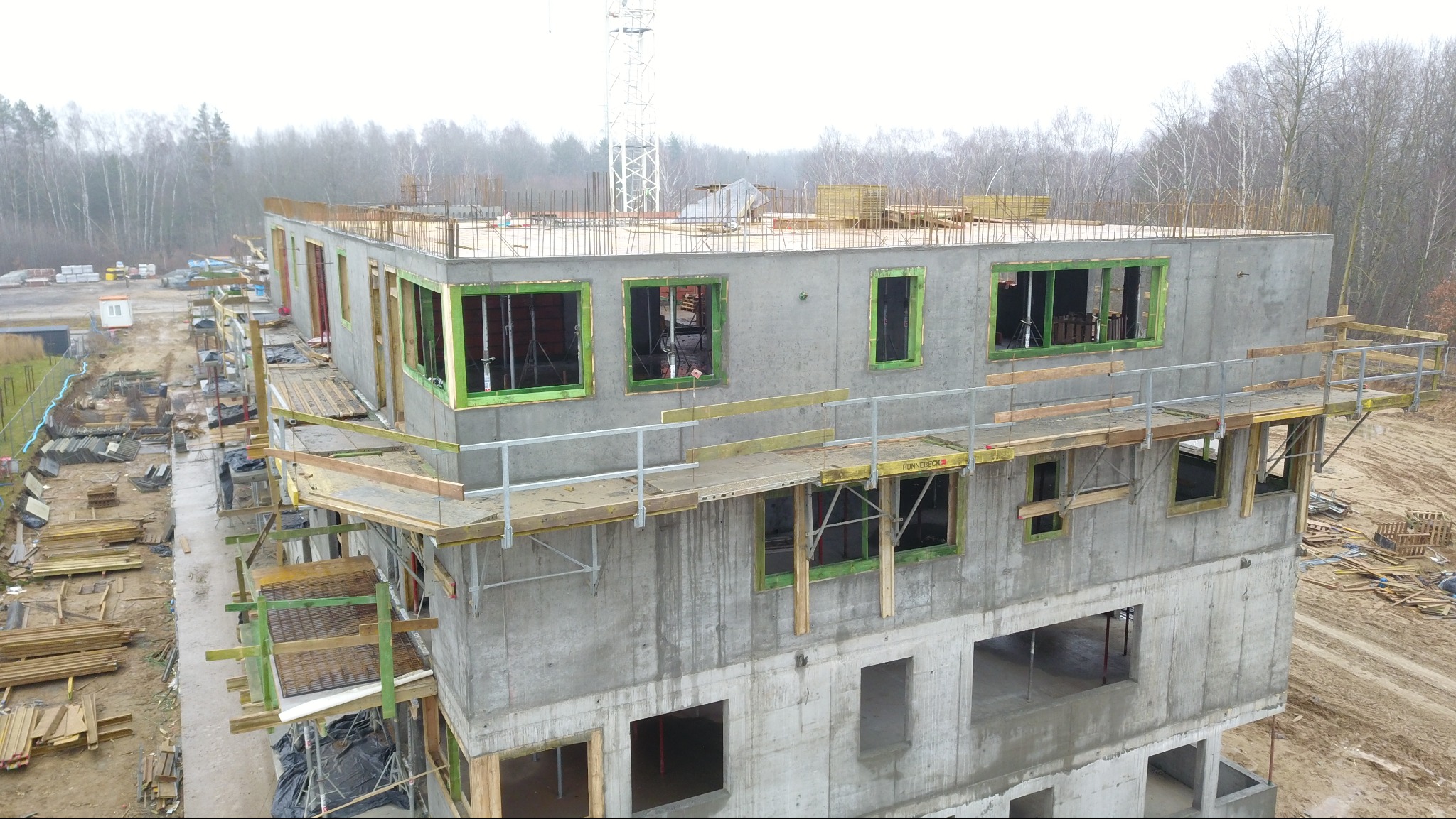 Surowy budynek żelbetowy w trakcie budowy, z widocznymi oknami zabezpieczonymi folią i szalunkami, rusztowaniami oraz zbrojeniami na dachu. Widok z góry.