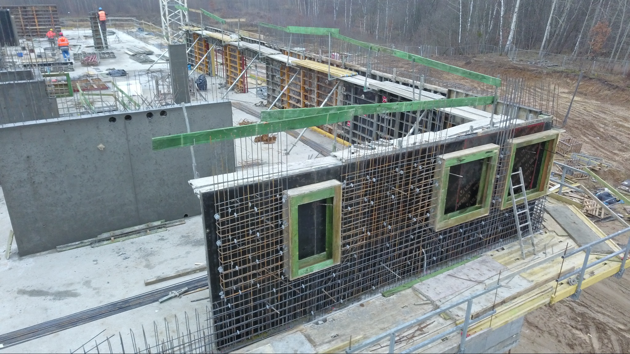 Budowa budynku żelbetowego: szalunek zbrojony z otworami okiennymi, widok z góry na plac budowy z pracownikami w oddali. Szare, pochmurne niebo w tle.