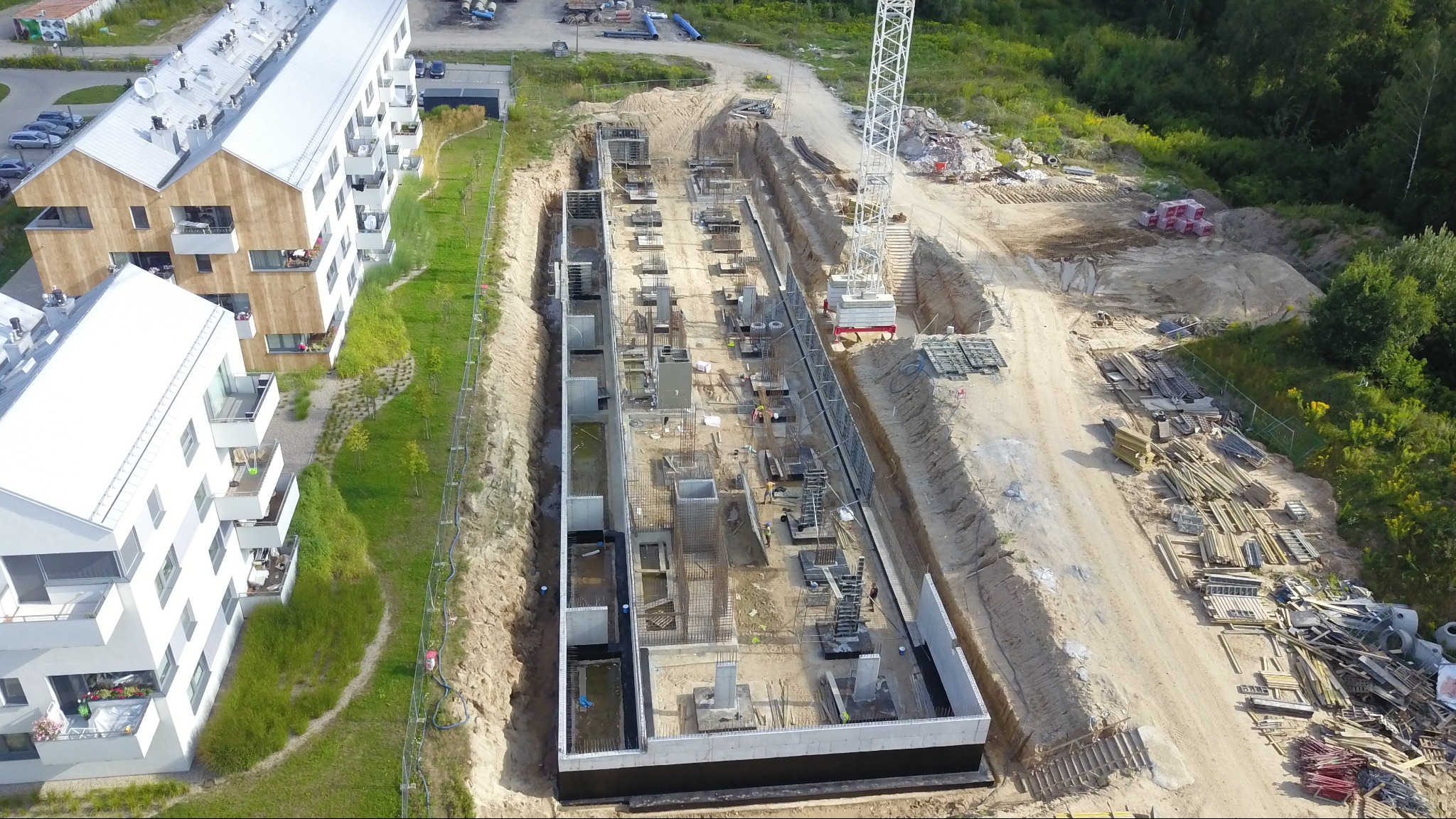 Zdjęcie z drona: fundamenty budynku wielorodzinnego z izolacją, zbrojeniami i szalunkami. Widoczny dźwig i teren budowy obok istniejących budynków.