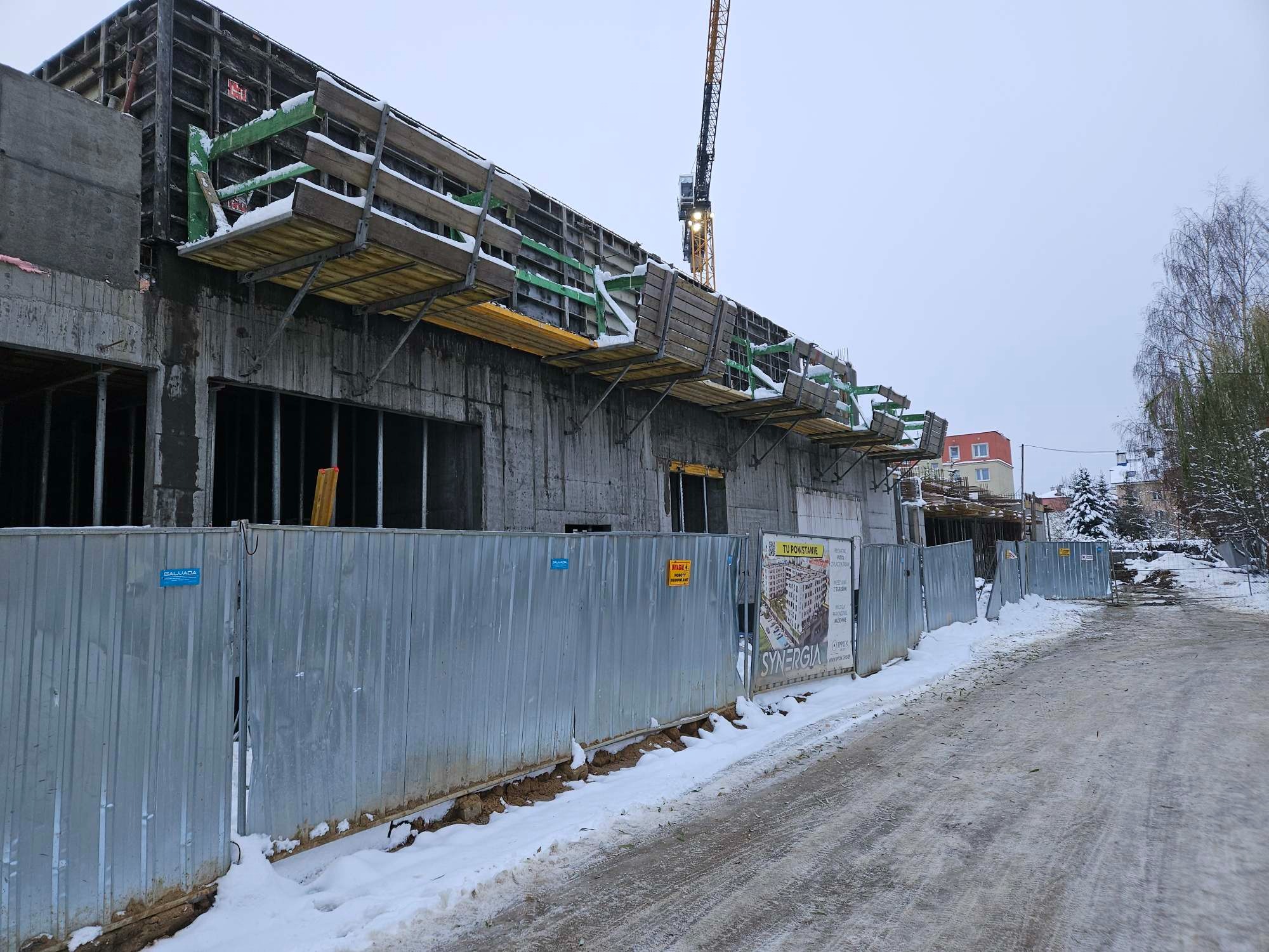 Budowa budynku żelbetowego w zimowej scenerii. Widoczne szalunki, rusztowania i dźwig. Teren budowy zabezpieczony metalowym ogrodzeniem z reklamą inwestycji.