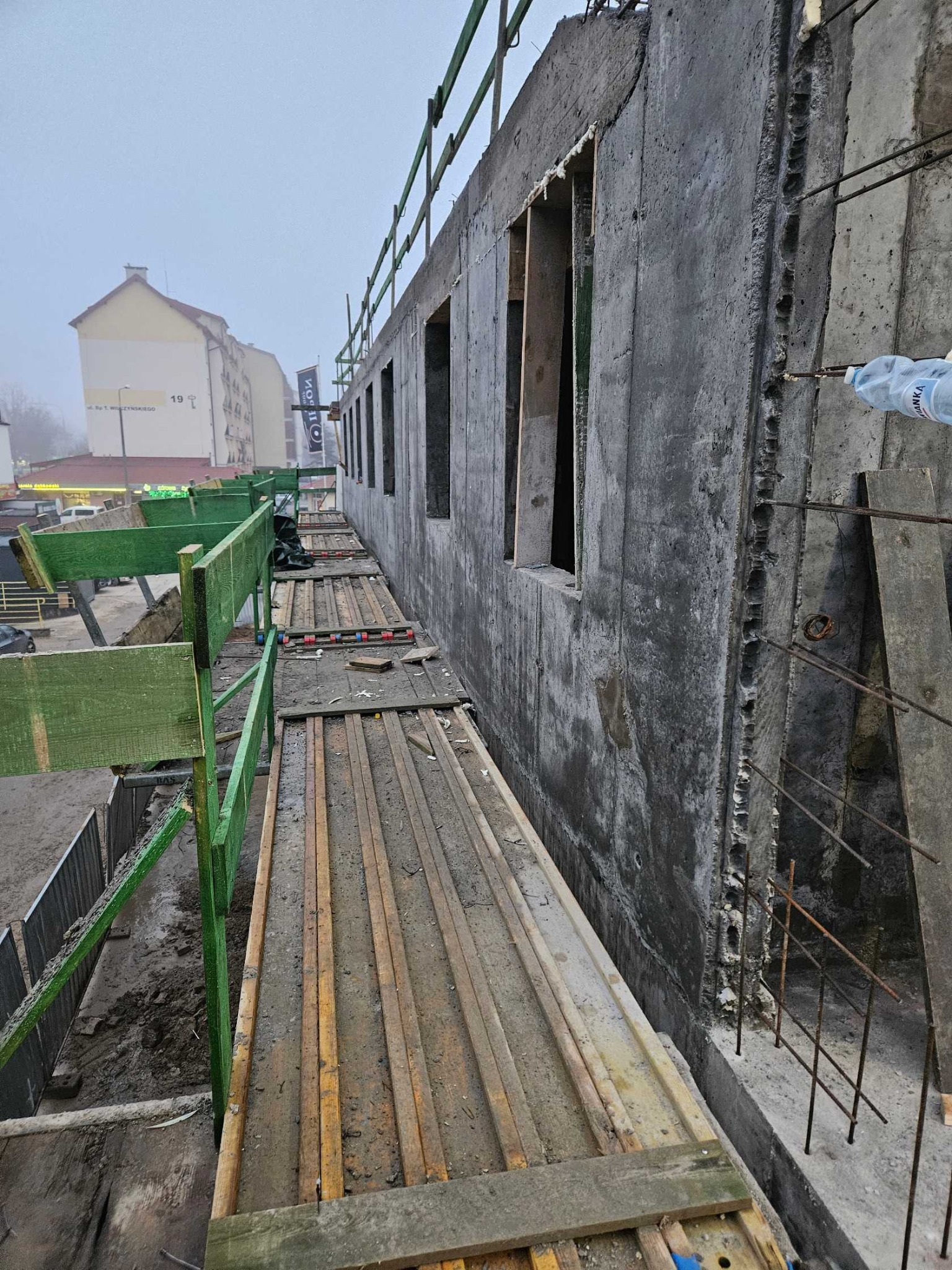 Surowa ściana żelbetowa z otworami okiennymi, widoczna z poziomu rusztowania. Szalunki i zbrojenia w tle, budynek w oddali, pochmurna pogoda.