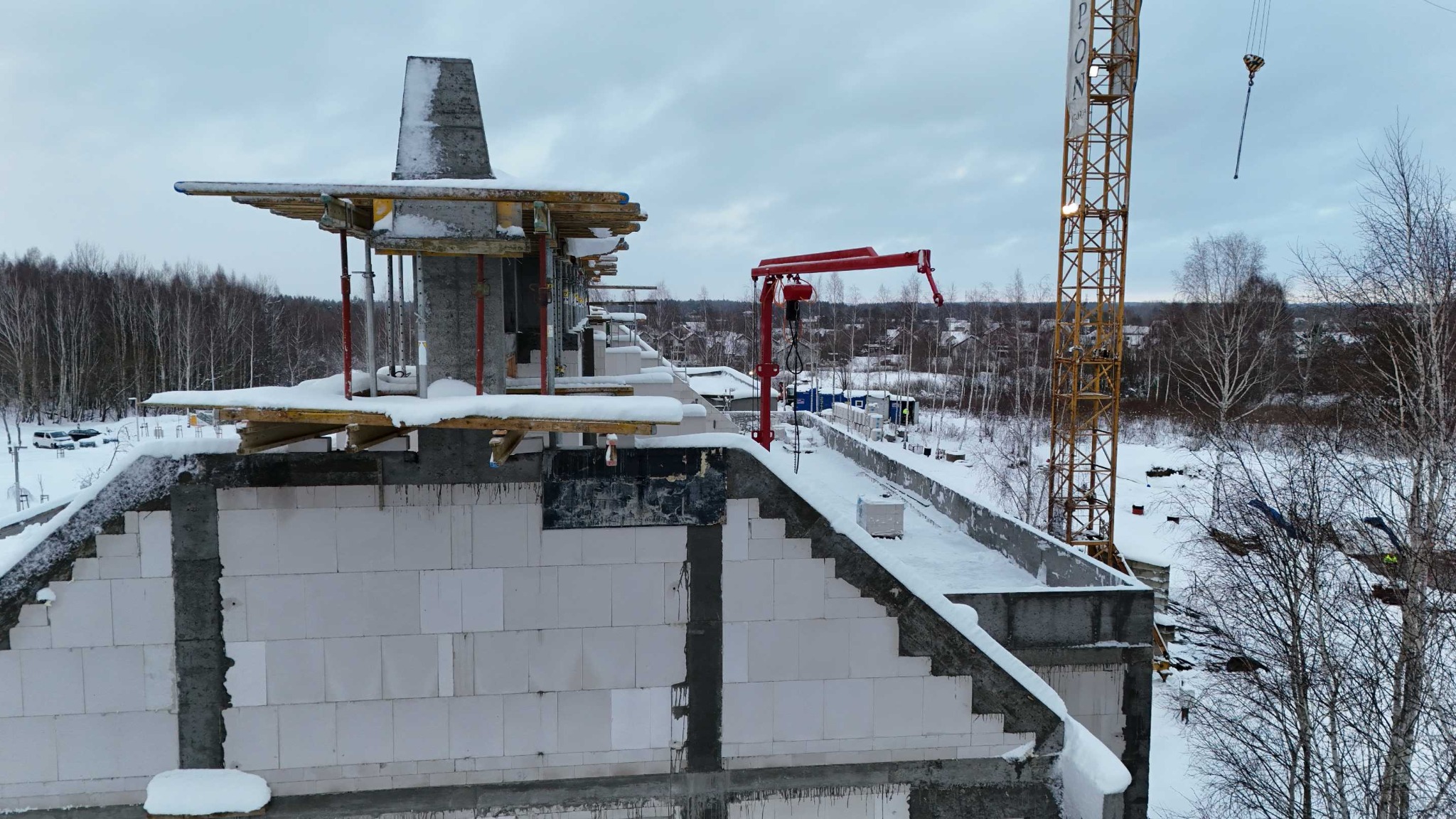 Budowa domu z betonu komórkowego zimą, widok z góry. Śnieg na elementach konstrukcyjnych, w tle las i dźwig budowlany. Widoczne szalunki i zbrojenia.
