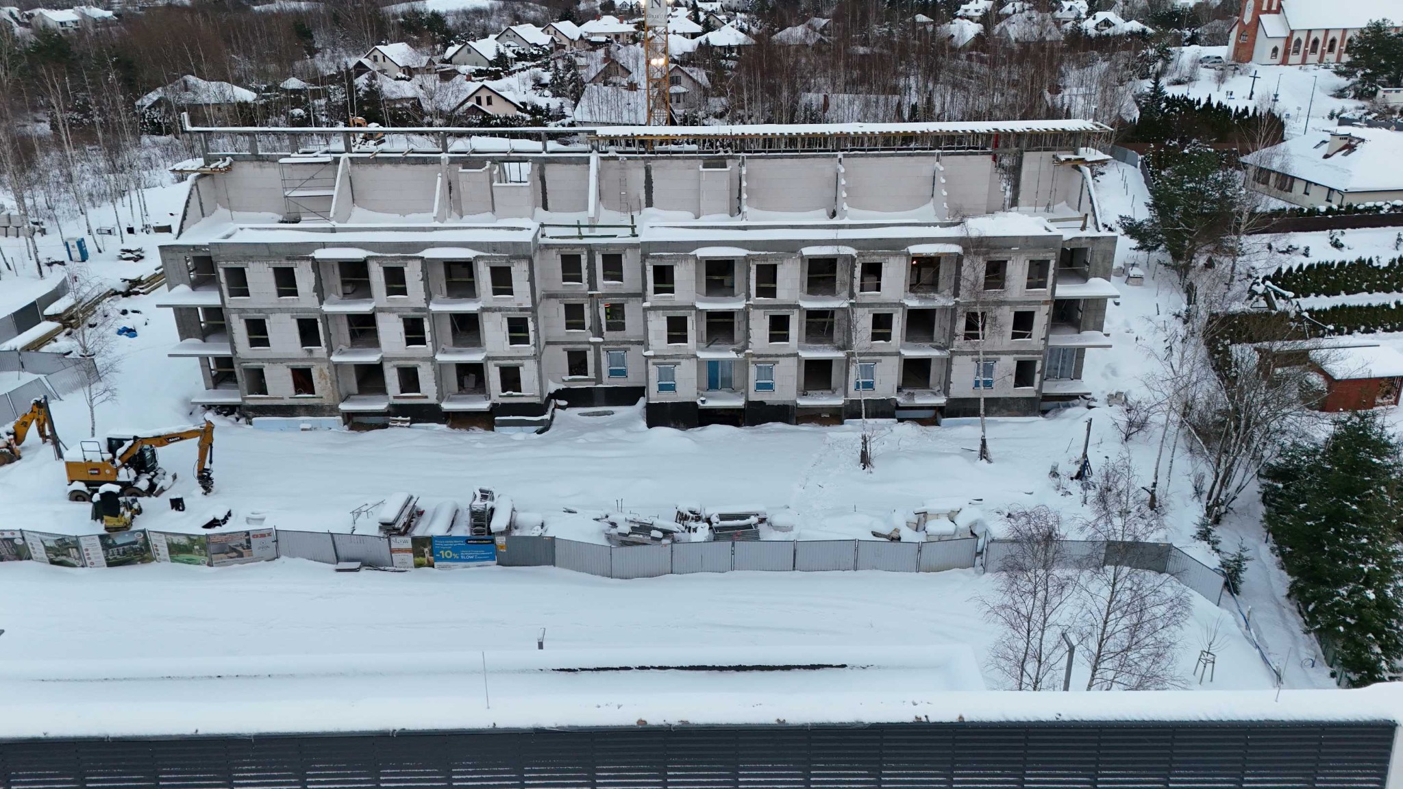 Wysoki budynek w stanie surowym zimą, z widocznym żurawiem i sprzętem budowlanym. Śnieżny krajobraz i zabudowa w tle. Ujęcie z góry.