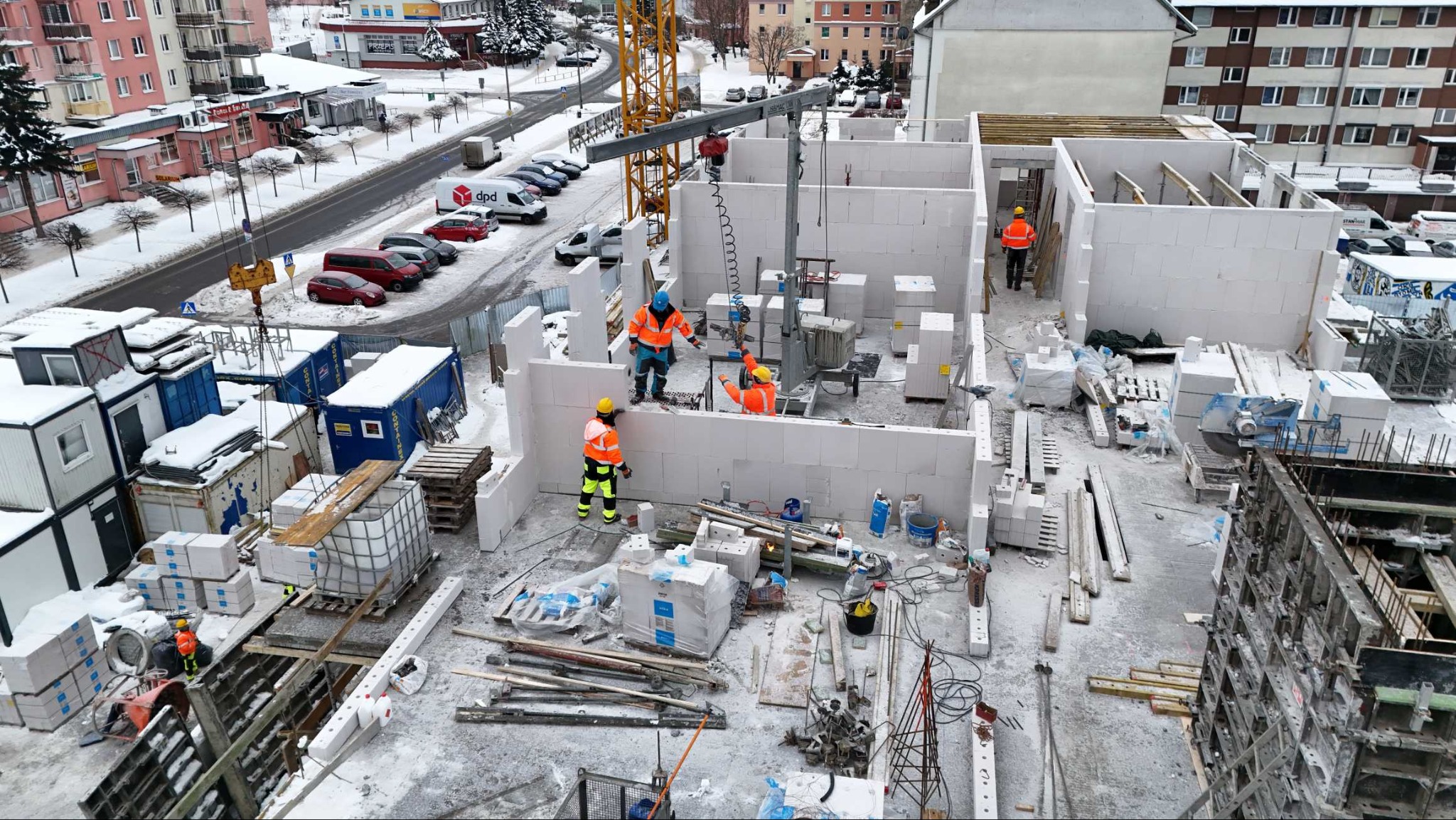 Budowa domu w zimowej scenerii: murarze w pomarańczowych kurtkach układają białe bloczki, używając dźwigu. Widok z góry na plac budowy i okoliczne budynki.