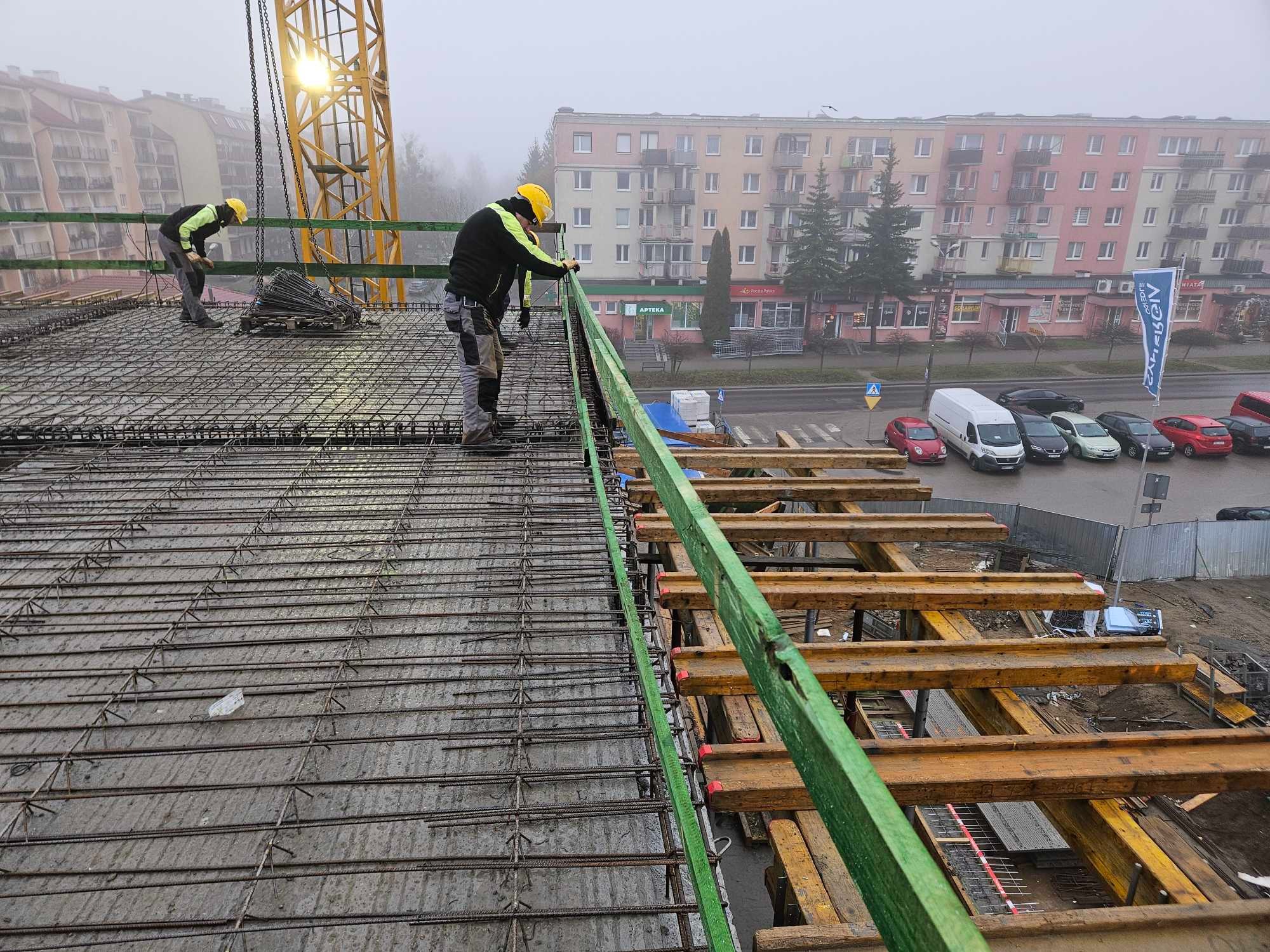 Zbrojenie stropu budynku w trakcie budowy, widoczne elementy szalunku i pracujący zbrojarze w kaskach, w tle budynki mieszkalne i pochmurne niebo.