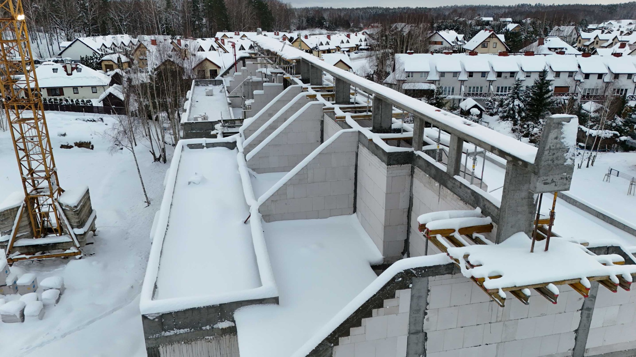 Budowa domu w zimowej scenerii: ściany z betonu komórkowego, szalunki, dźwig w tle, wszystko pokryte śniegiem. Widok z góry na osiedle domów.
