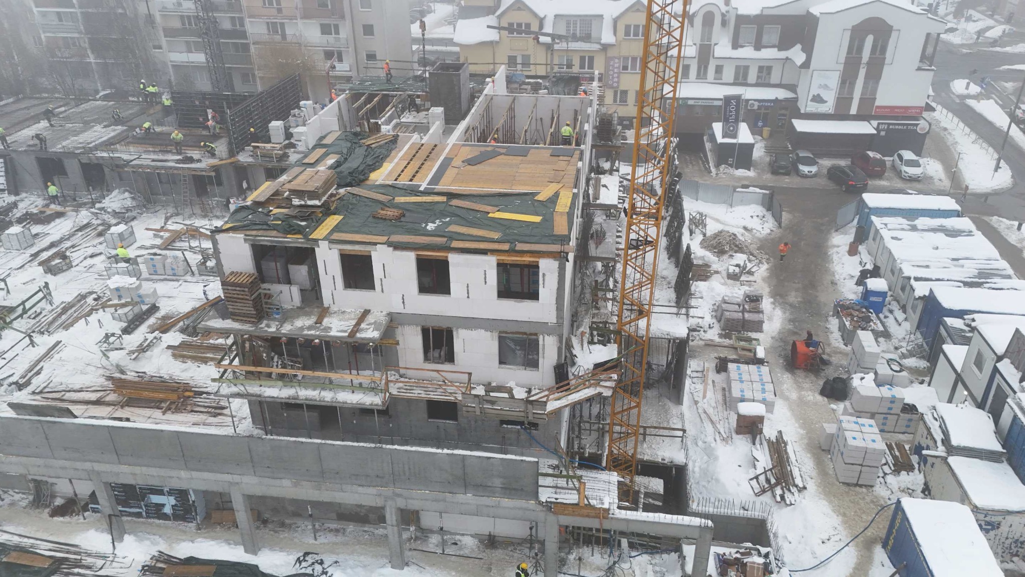 Wysoki kąt na budowę w zimie: dwukondygnacyjny budynek z białych bloków, częściowo przykryty plandeką, z żurawiem budowlanym obok. Śnieg na placu budowy.