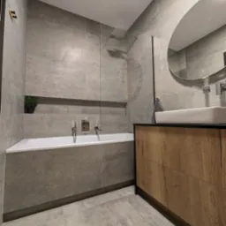 Nowoczesna łazienka z szarymi płytkami, wanną zabudowaną płytkami, umywalką nablatową na szafce z drewnianym frontem i owalnym lustrem.