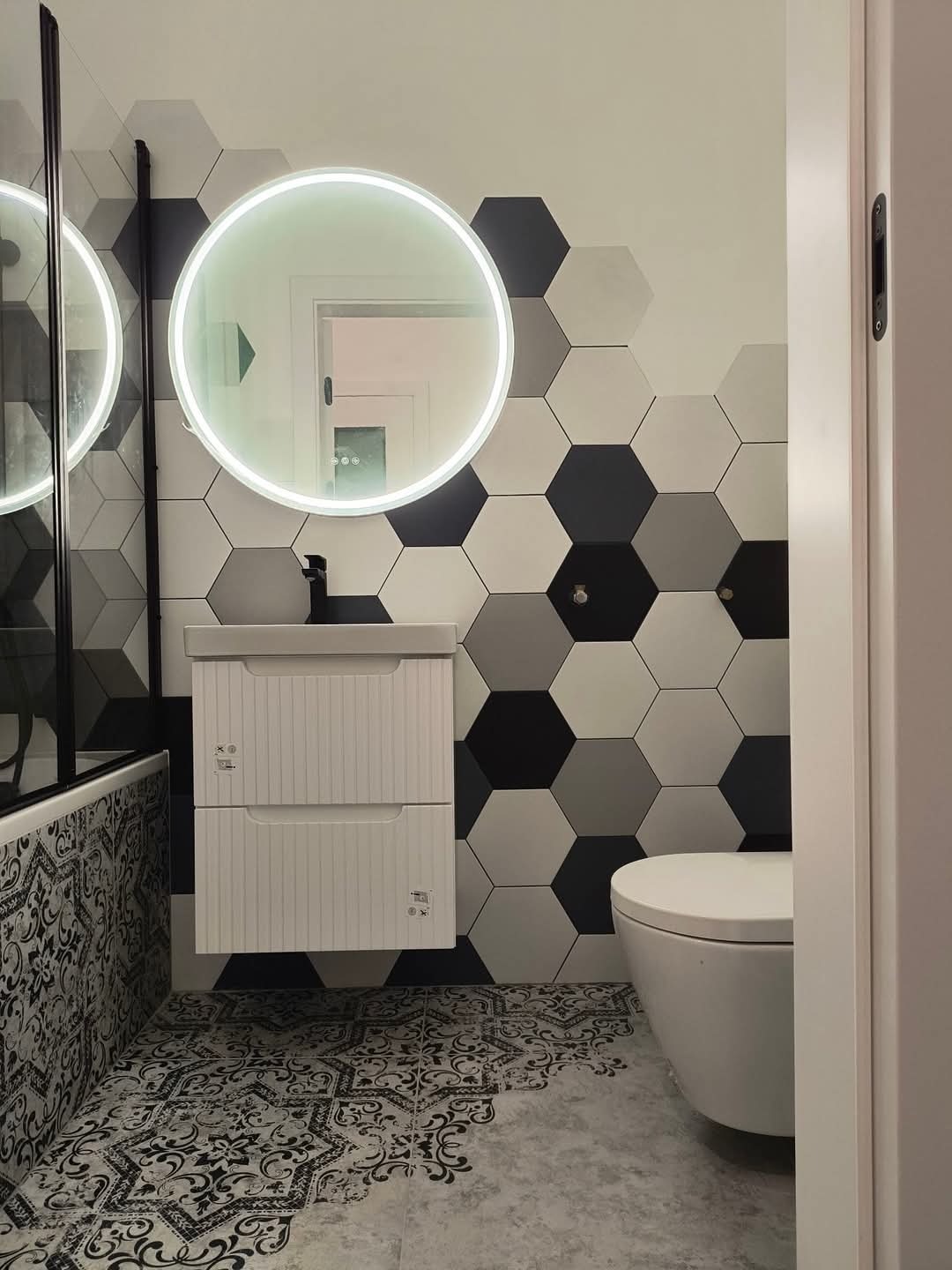 Wykończona łazienka z heksagonalnymi płytkami w odcieniach szarości na ścianie, okrągłym lustrem z podświetleniem LED, białą szafką pod umywalką i wzorzystą płytką podłogową.