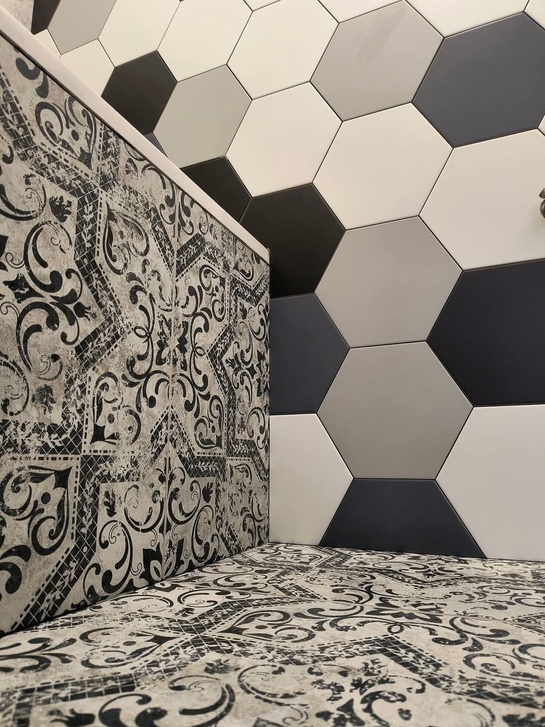 Ściana i podłoga wyłożone płytkami w geometryczne wzory: heksagonalne płytki w odcieniach szarości i białe na ścianie oraz płytki z ornamentem w czerni i bieli na podłodze i ścianie