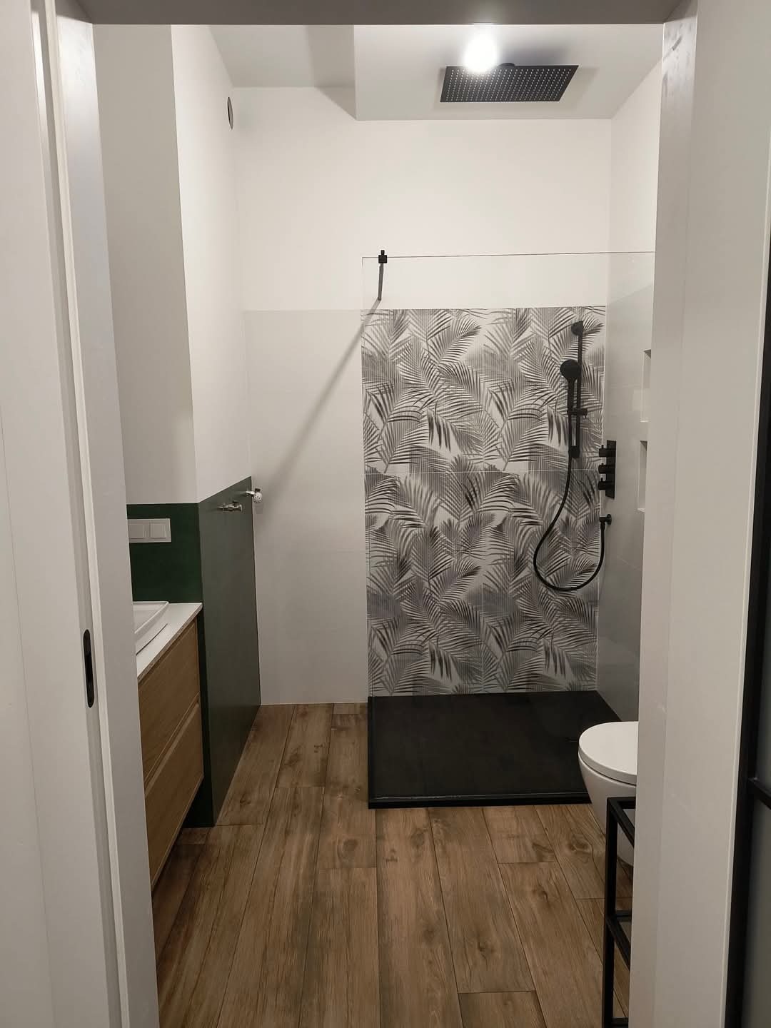 Nowoczesna łazienka z kabiną prysznicową typu walk-in, wyłożoną płytkami w motywie liści palmowych, czarną armaturą i drewnopodobną podłogą, widok zza otwartych drzwi.