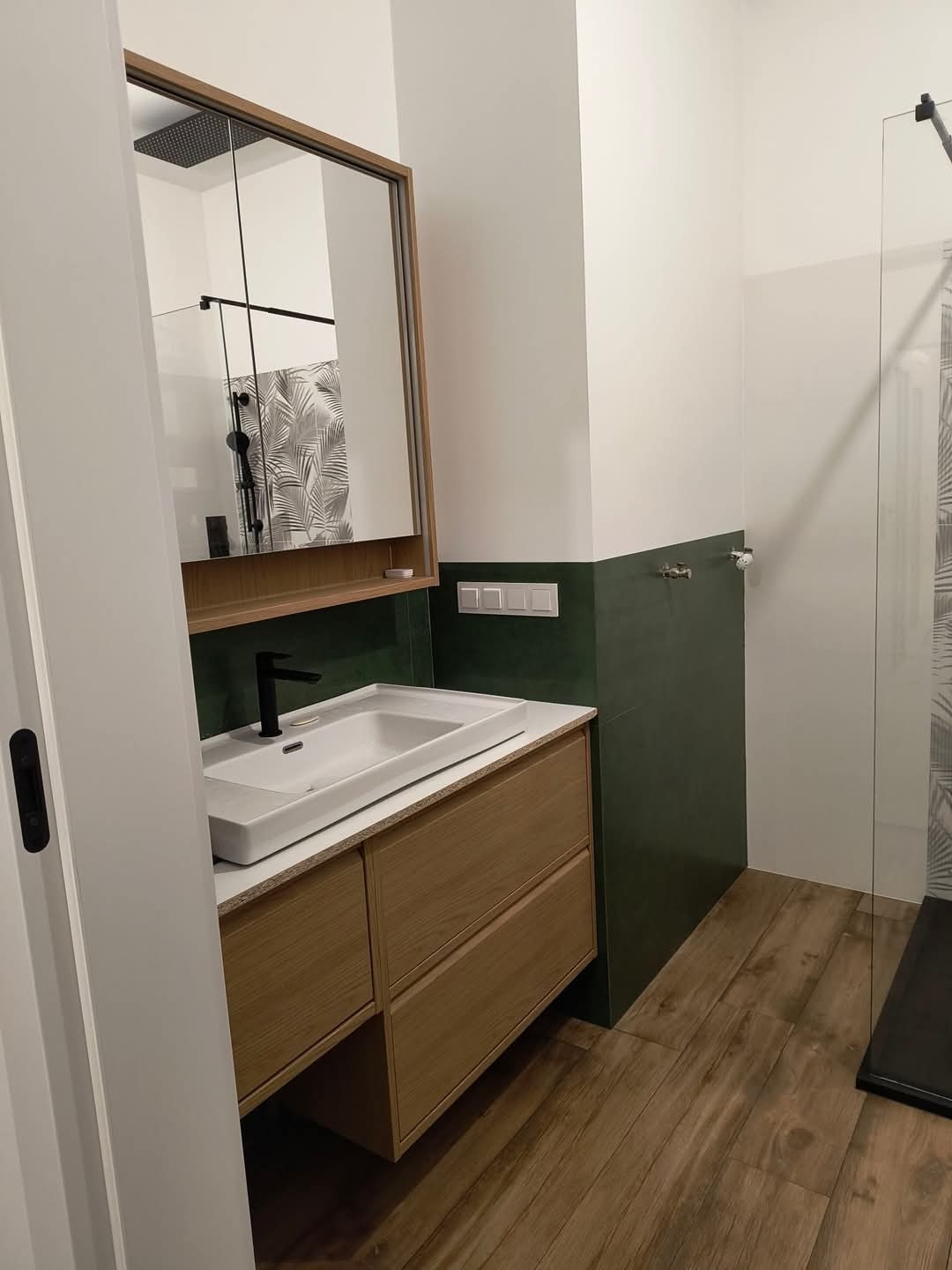 Nowoczesna łazienka z drewnianą szafką pod umywalką, czarną armaturą i zielonymi akcentami na ścianach, widok z perspektywy otwartych drzwi.