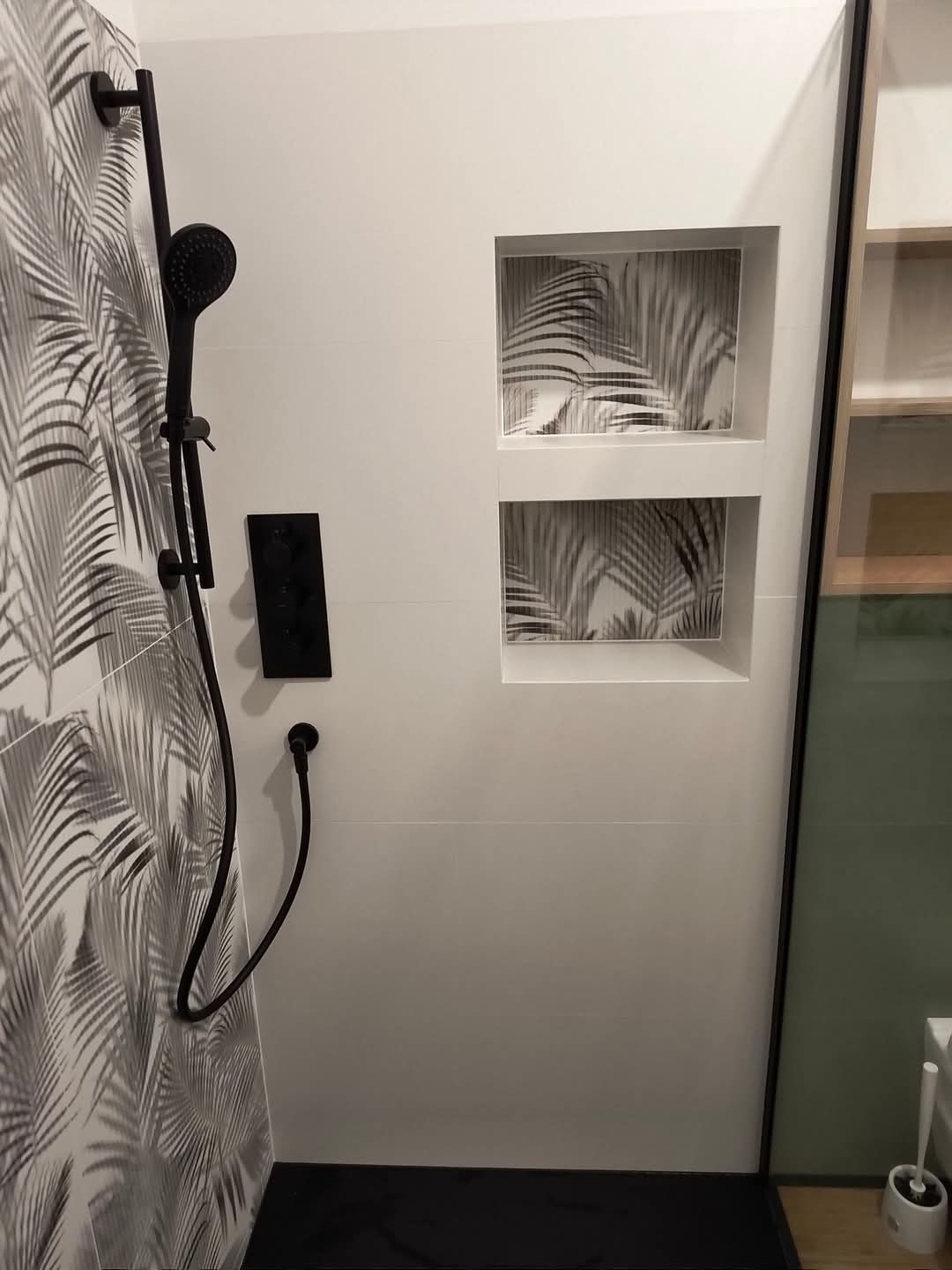 Nowoczesna kabina prysznicowa z czarną armaturą i płytkami w motywie liści palmowych, wnęki dekoracyjne z tym samym motywem, zrealizowana w Łomiankach dolnych.