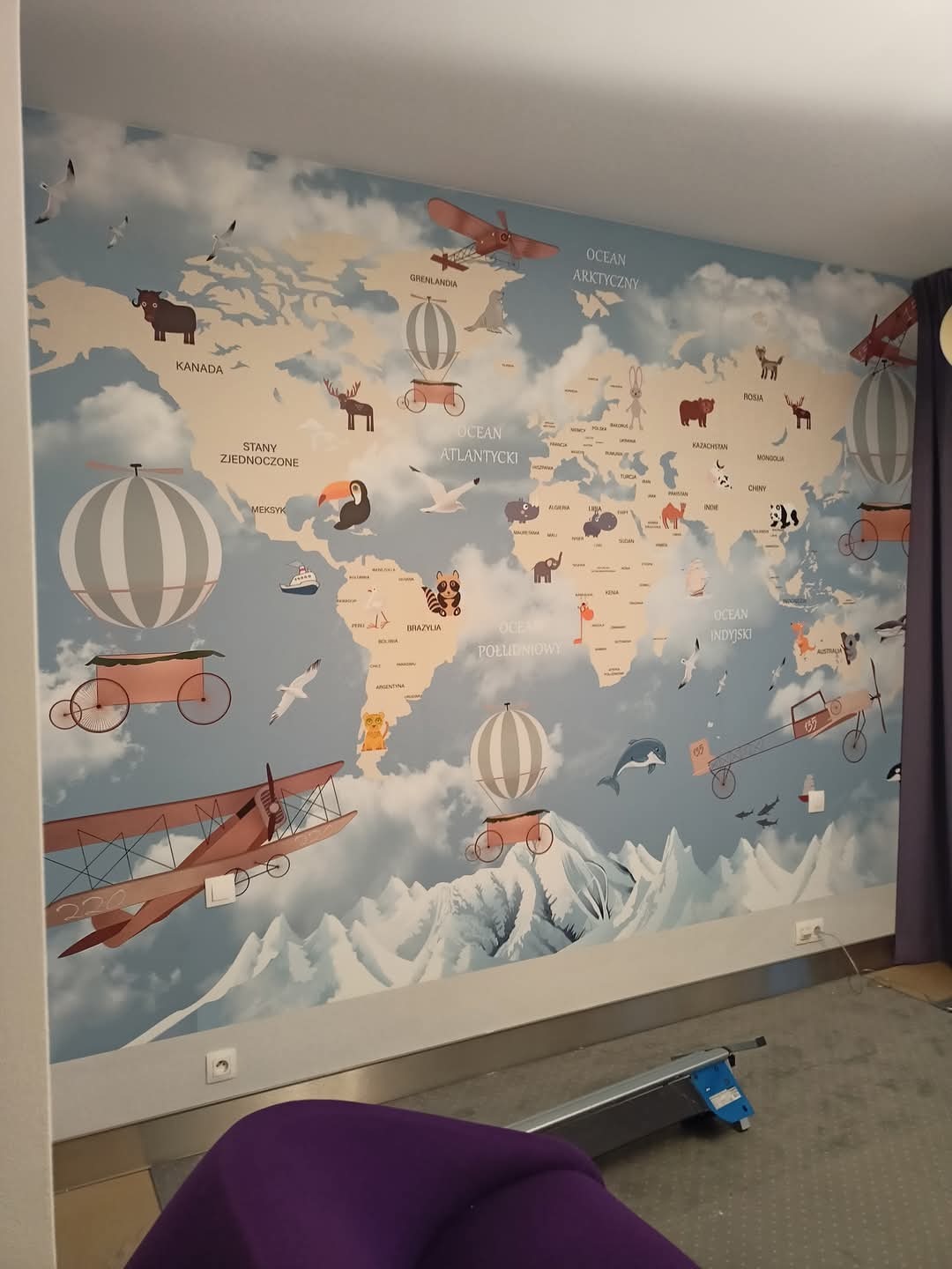 Tapeta na ścianie pokoju dziecięcego z motywem mapy świata, ozdobiona rysunkami zwierząt, samolotów i balonów, widoczne gniazdka elektryczne na ścianie.