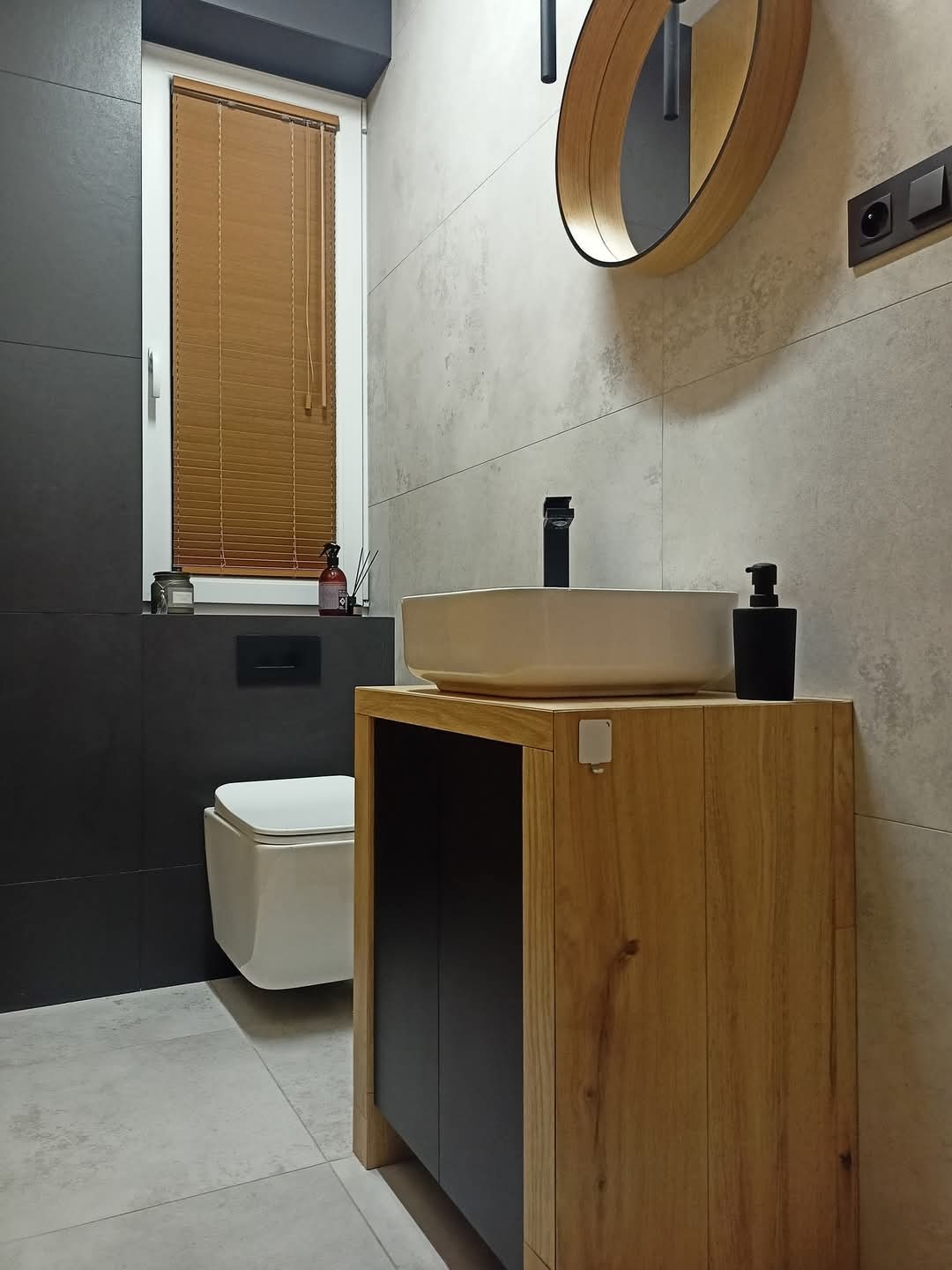 Nowoczesna łazienka z wiszącą toaletą, umywalką nablatową na drewnianej szafce z czarnymi frontami, okrągłym lustrem i brązowymi żaluzjami w oknie.