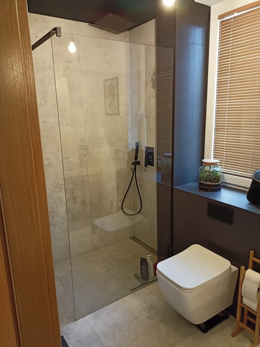 Nowoczesna łazienka z kabiną prysznicową typu walk-in, szare płytki imitujące beton, deszczownica, wisząca miska WC i drewniane żaluzje w oknie.