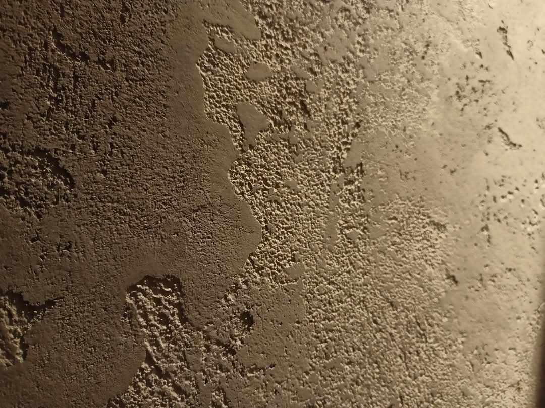 Fragment ściany z dekoracyjną, strukturalną powierzchnią w odcieniach beżu i brązu, oświetlony z boku, ukazujący nieregularne wzory i fakturę.