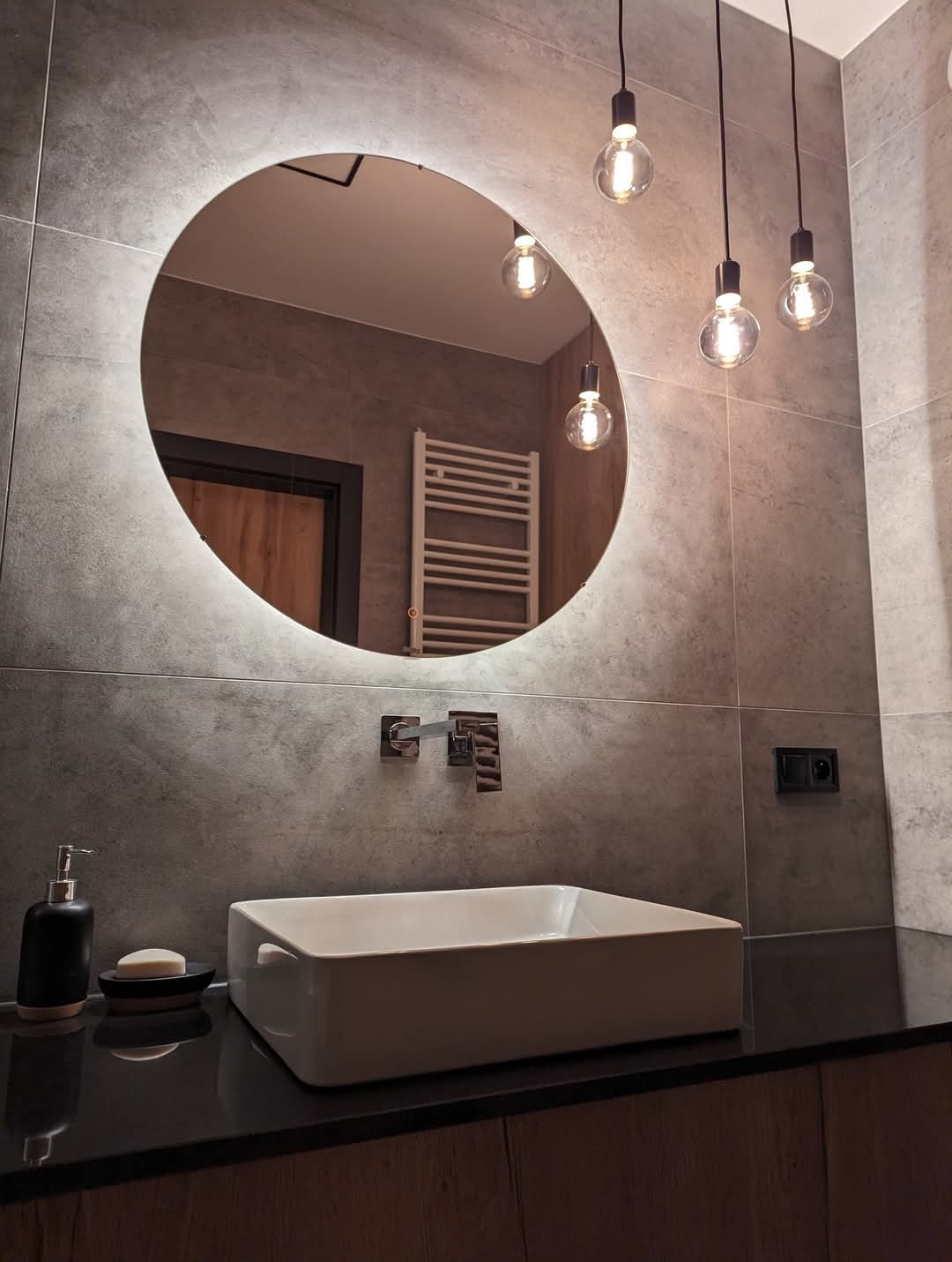 Nowoczesna łazienka z owalnym lustrem podświetlanym LED, wiszącymi żarówkami Edisona, prostokątną umywalką nablatową i szarymi płytkami imitującymi beton.