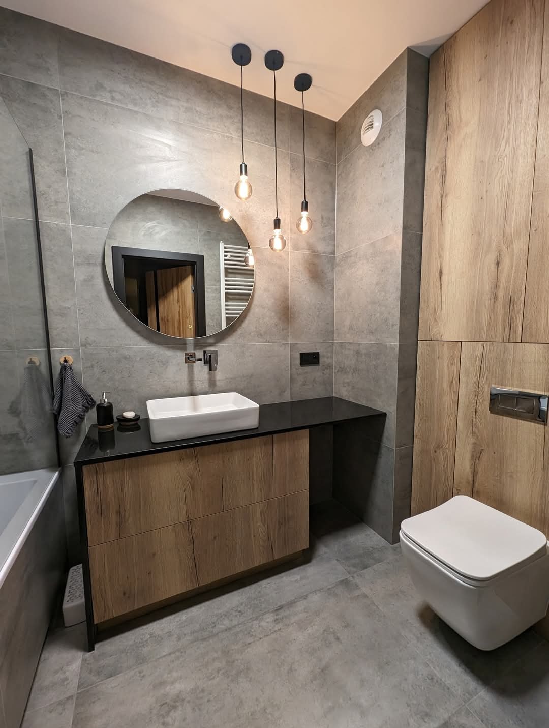 Nowoczesna łazienka z szarymi płytkami, drewnianymi akcentami, okrągłym lustrem i wiszącymi lampami z żarówkami Edisona, z widoczną toaletą bezkołnierzową i czarnym blatem umywalkowym.