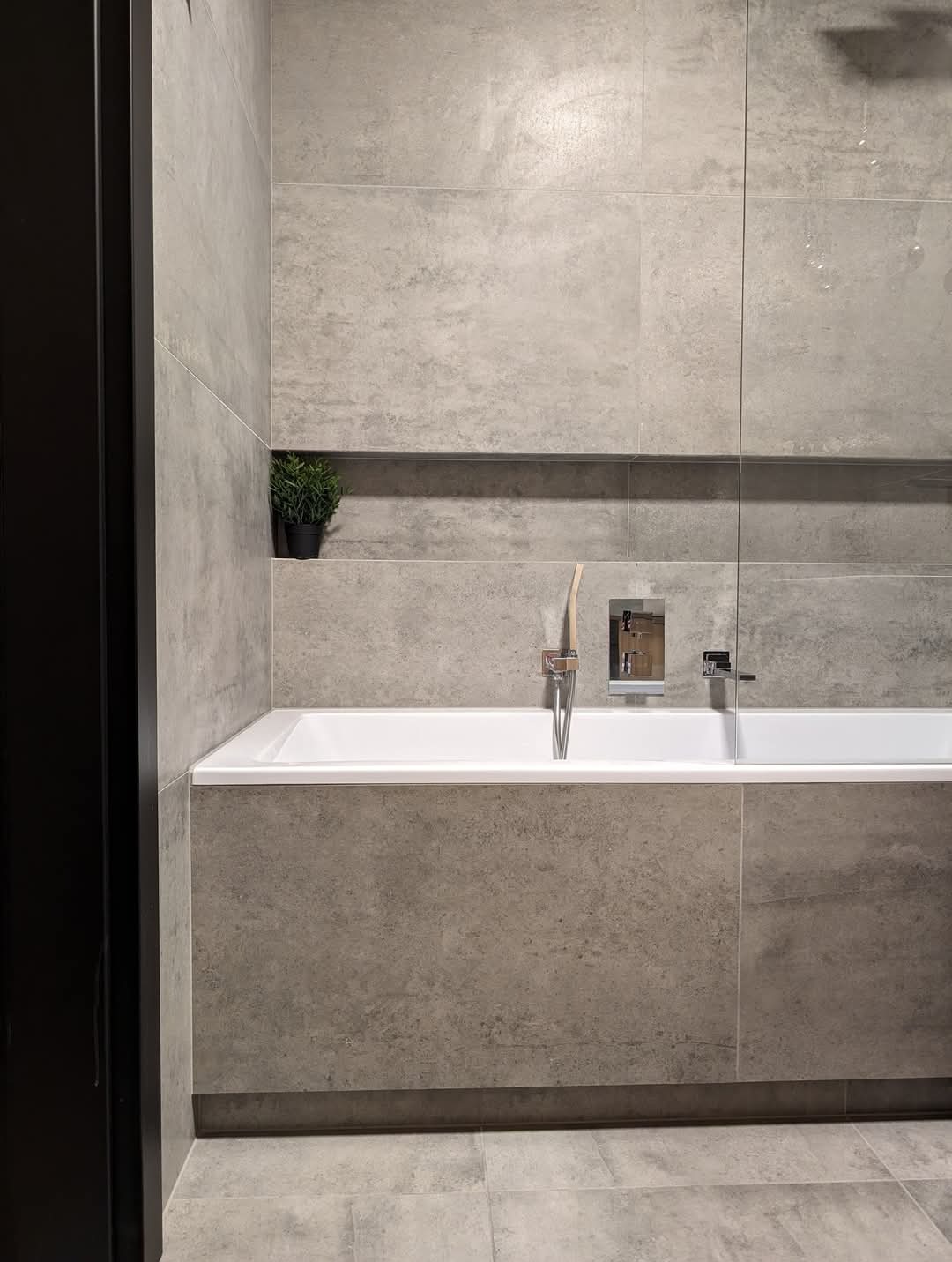 Minimalistyczna łazienka w odcieniach szarości z wanną obudowaną płytkami, chromowaną armaturą i małą rośliną doniczkową w niszy ściennej.