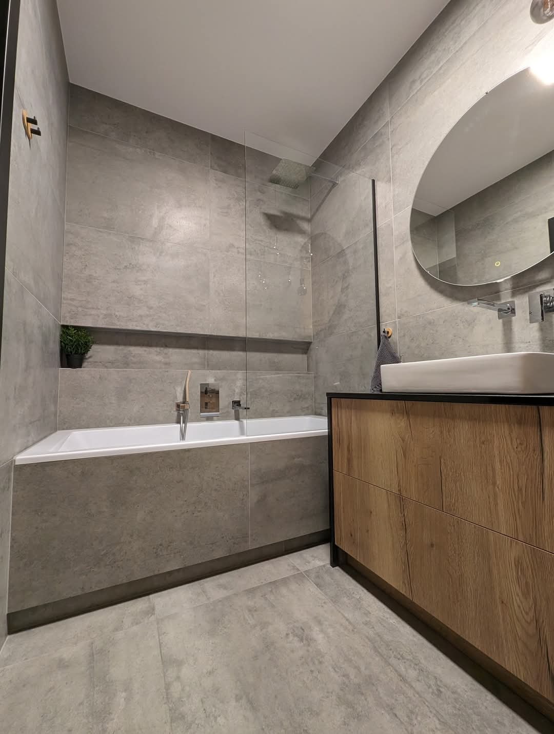 Nowoczesna łazienka z szarymi płytkami, wanną zabudowaną płytkami, umywalką nablatową na szafce z drewnianym frontem i owalnym lustrem.