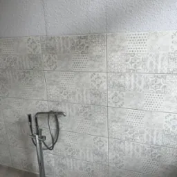 Wykończona ściana łazienki z dekoracyjnymi płytkami w odcieniach szarości, z widoczną chromowaną baterią wannową wolnostojącą, oraz fragmentem ściany pokrytej gładzią strukturalną.