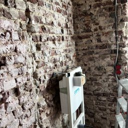 Builder Atta - Montaż stelaża podtynkowego WC na tle surowej ściany z cegły, widoczne przewody elektryczne i drabina.