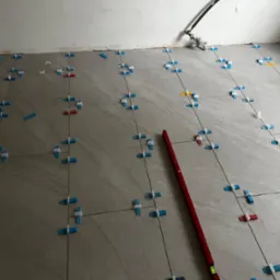 Układanie szarych płytek podłogowych z użyciem niebieskich krzyżyk&oacute;w dystansowych i czerwonej poziomicy, widoczne wystające kable ze ściany.