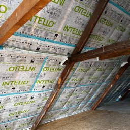 Builder Atta - Poddasze z widoczną izolacją termiczną marki Intelio, zamontowaną między krokwiami, zabezpieczoną taśmą uszczelniającą, widoczne drewniane elementy konstrukcji dachu i fragment podłogi z płyty OSB.