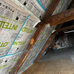 Montaż membrany paroizolacyjnej Intello na drewnianej konstrukcji poddasza, widoczne krokwie i płyty OSB, taśma uszczelniająca na łączeniach, w tle drabiny i elementy wyposażenia.
