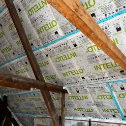 Builder Atta - Widok poddasza z widoczną membraną paroizolacyjną Intello, zamontowaną między krokwiami drewnianymi, z widocznymi napisami i symbolami na membranie.