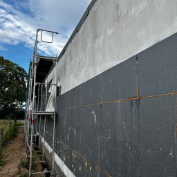 Builder Atta - Ocieplanie ściany zewnętrznej budynku styropianem grafitowym, widoczne żółte kołki mocujące, szpary wypełnione pianką montażową, rusztowanie przy ścianie.
